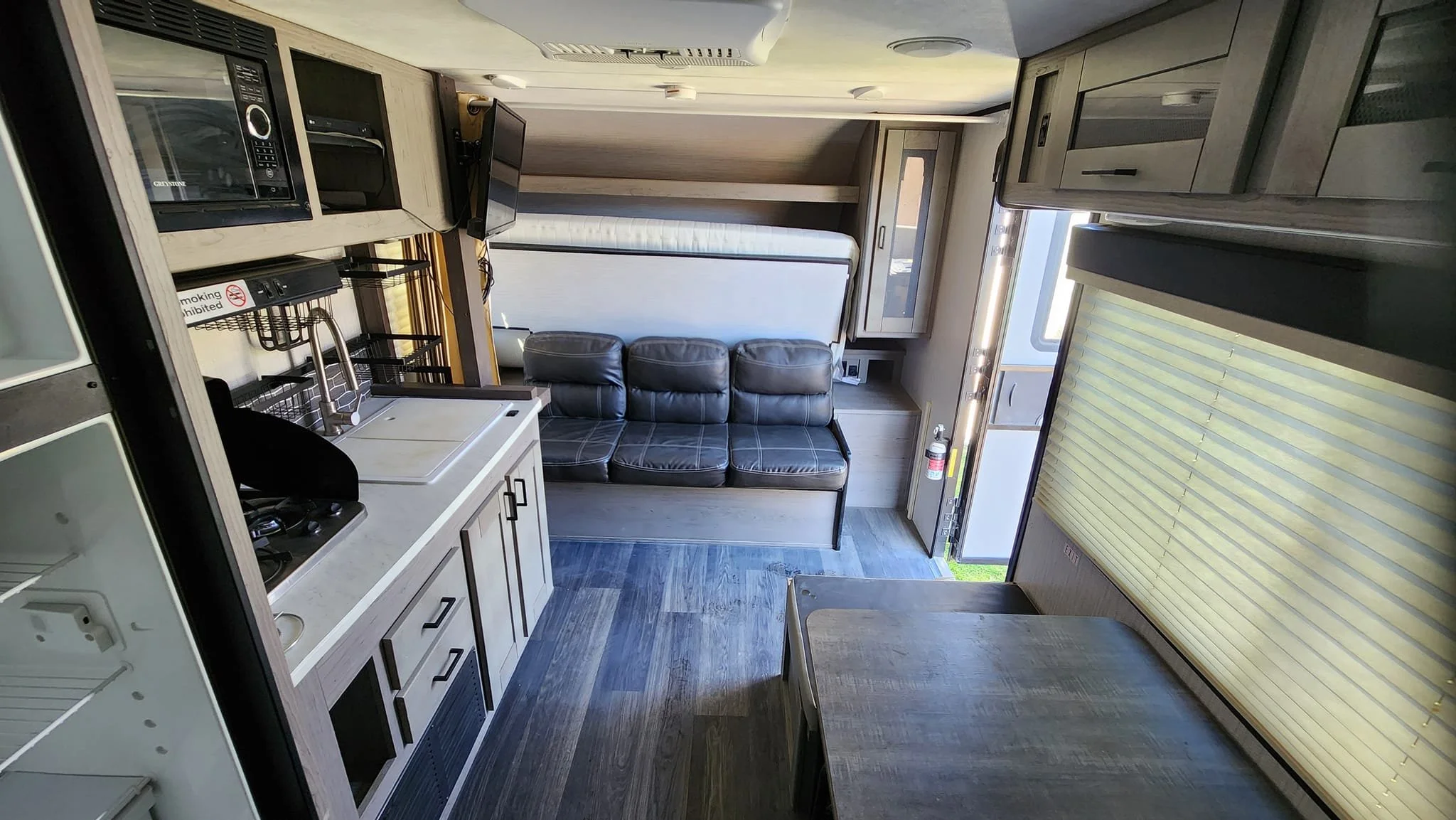Interior of a compact RV or camper with a kitchenette, seating area, and large window with blinds.