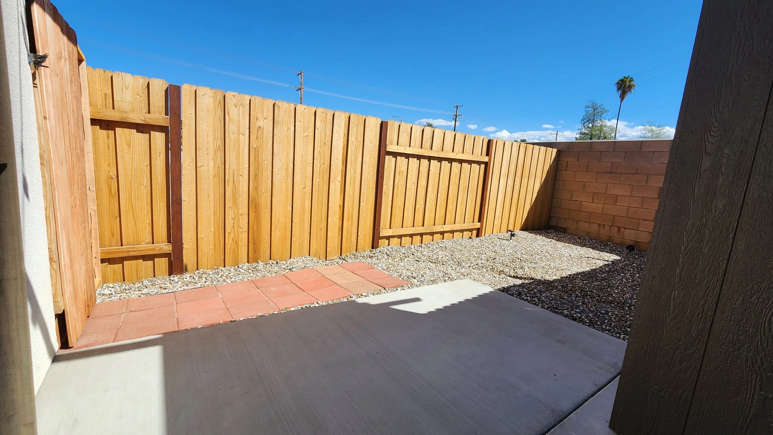 View of a backyard with a newly built wooden fence, small brick wall, gravel ground, and concrete patio under a bright blue sky.