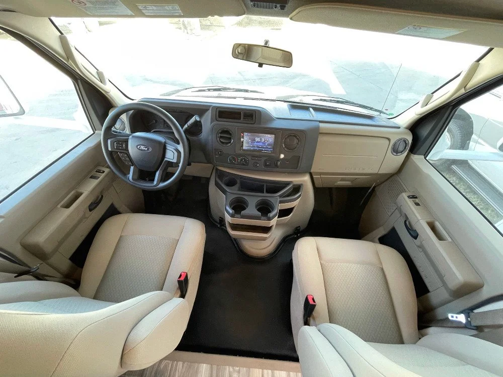 Interior view of a beige Ford RV or camper van cab with driver and passenger seats, dashboard, steering wheel, and center console.