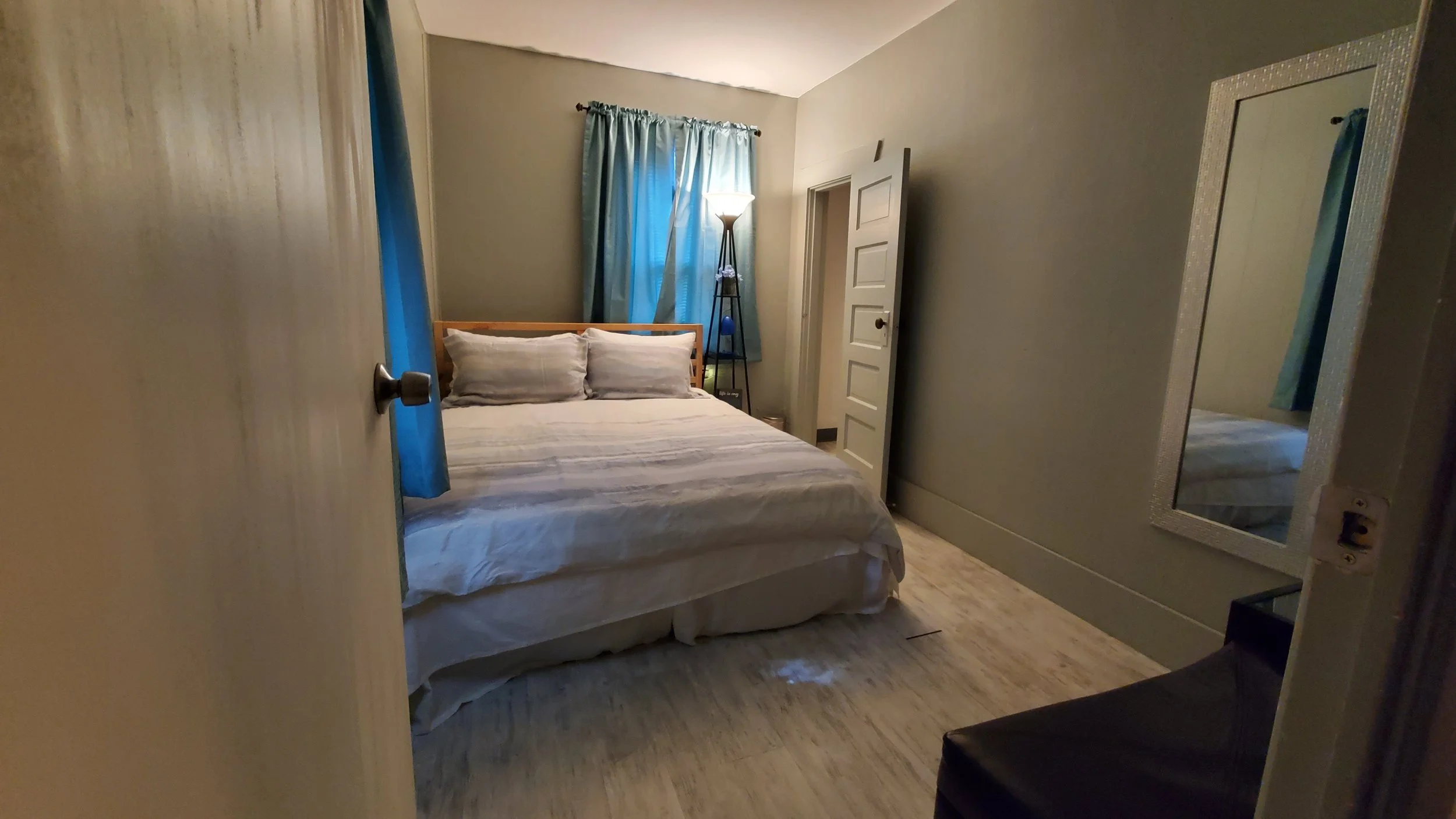 Photo of a small, cozy bedroom with a bed, blue curtains, a floor lamp, a mirror, and a dresser. The door is partially open, revealing the room inside.