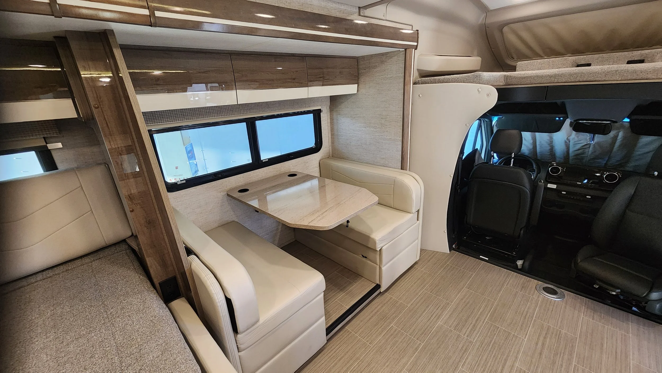 Interior of a recreational vehicle featuring a dining area with a beige booth and a marble table, a window with blinds, and the driver's area with seats and dashboard.
