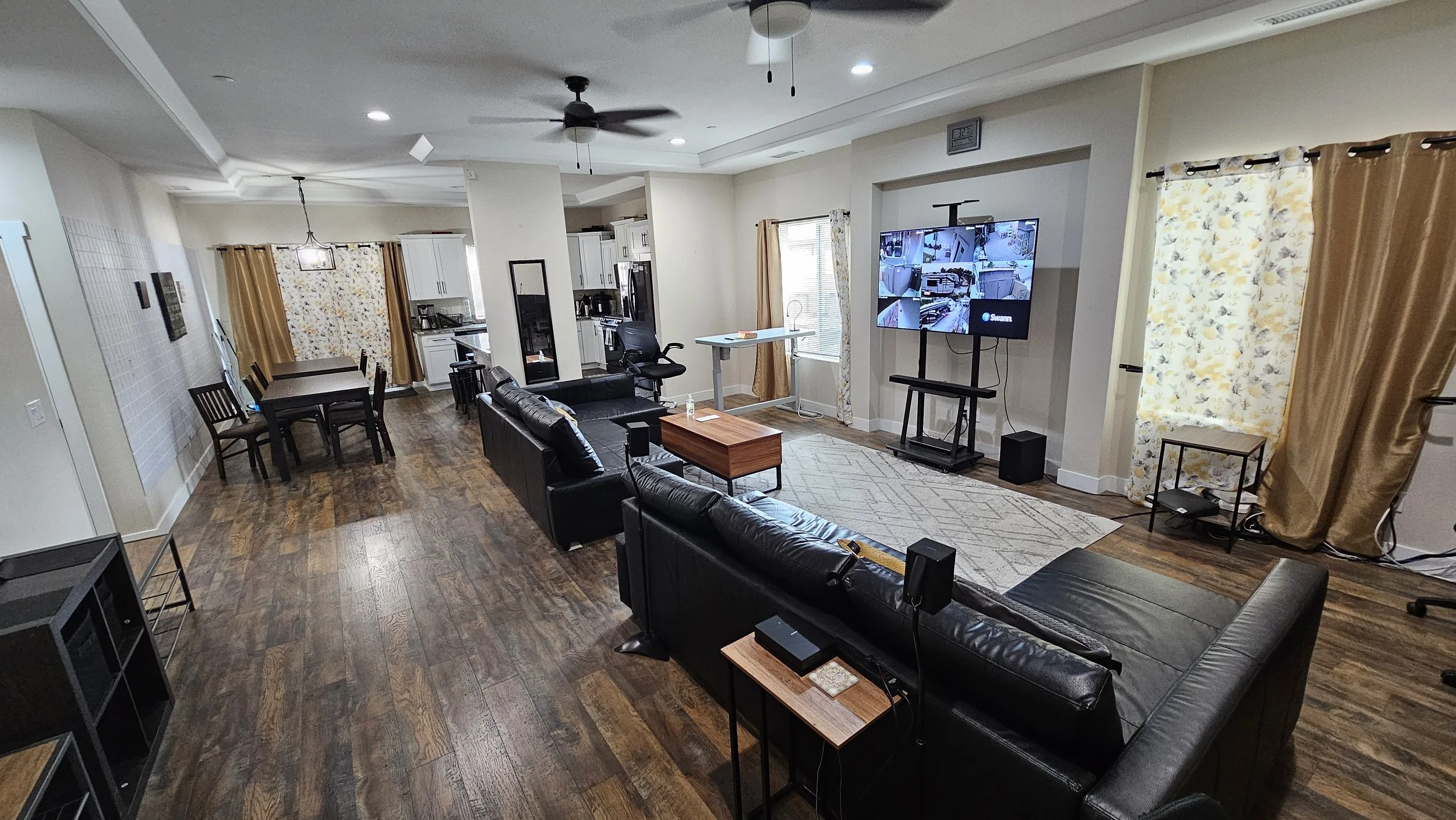 Open-concept living room and kitchen with hardwood floors, black leather sofas, a large wall-mounted TV, dining area, and kitchen with white cabinetry and stainless steel appliances, decorated with yellow and floral curtains.
