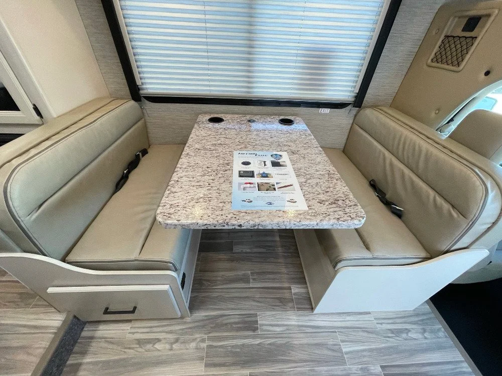 Booth-style seating with beige cushions around a granite tabletop inside an RV or camper, with a window and blinds in the background.