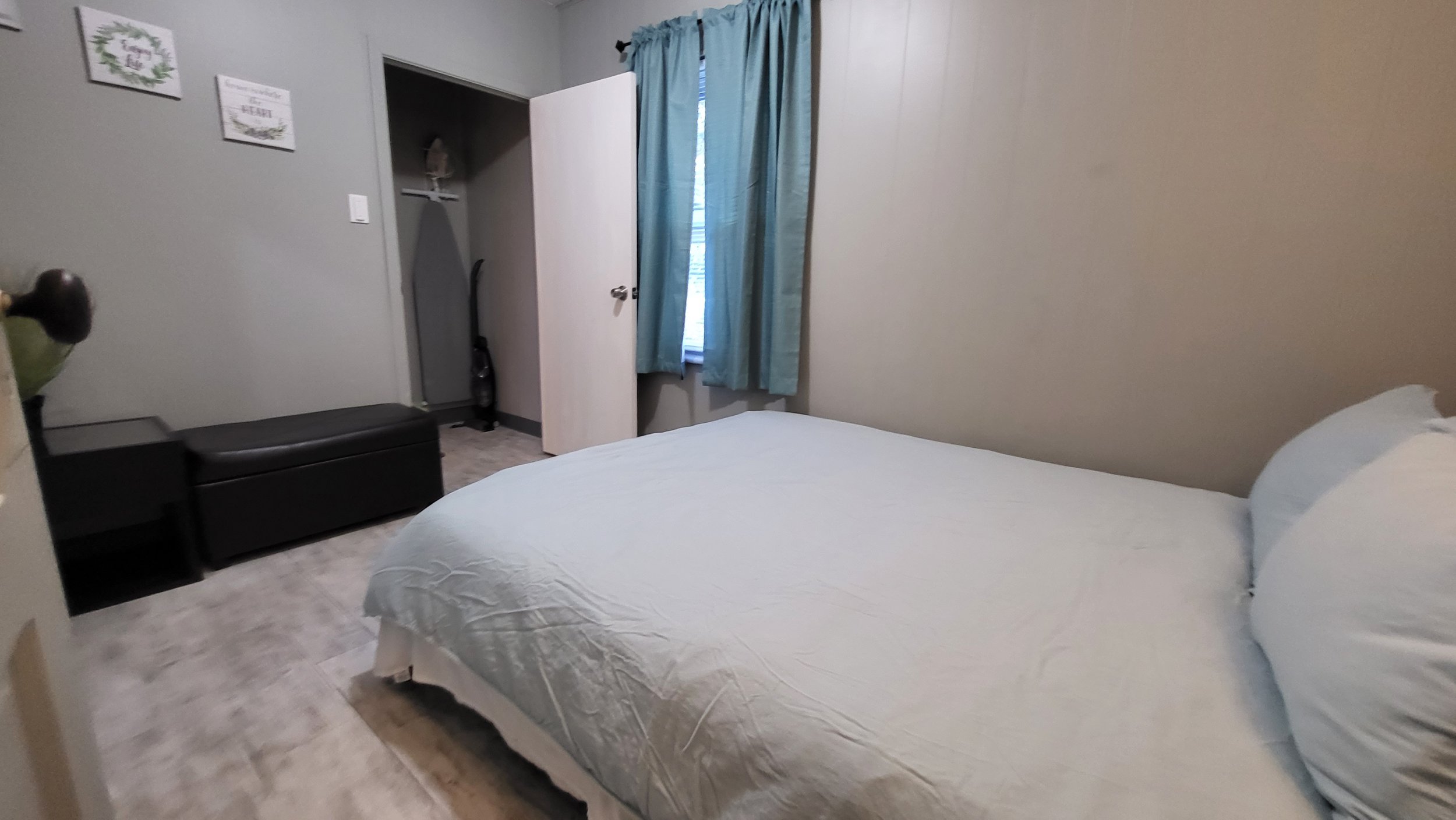 A bedroom with a bed, blue curtains, and a window with sunlight, a white wall, and a closet with an open door revealing a vacuum cleaner inside.