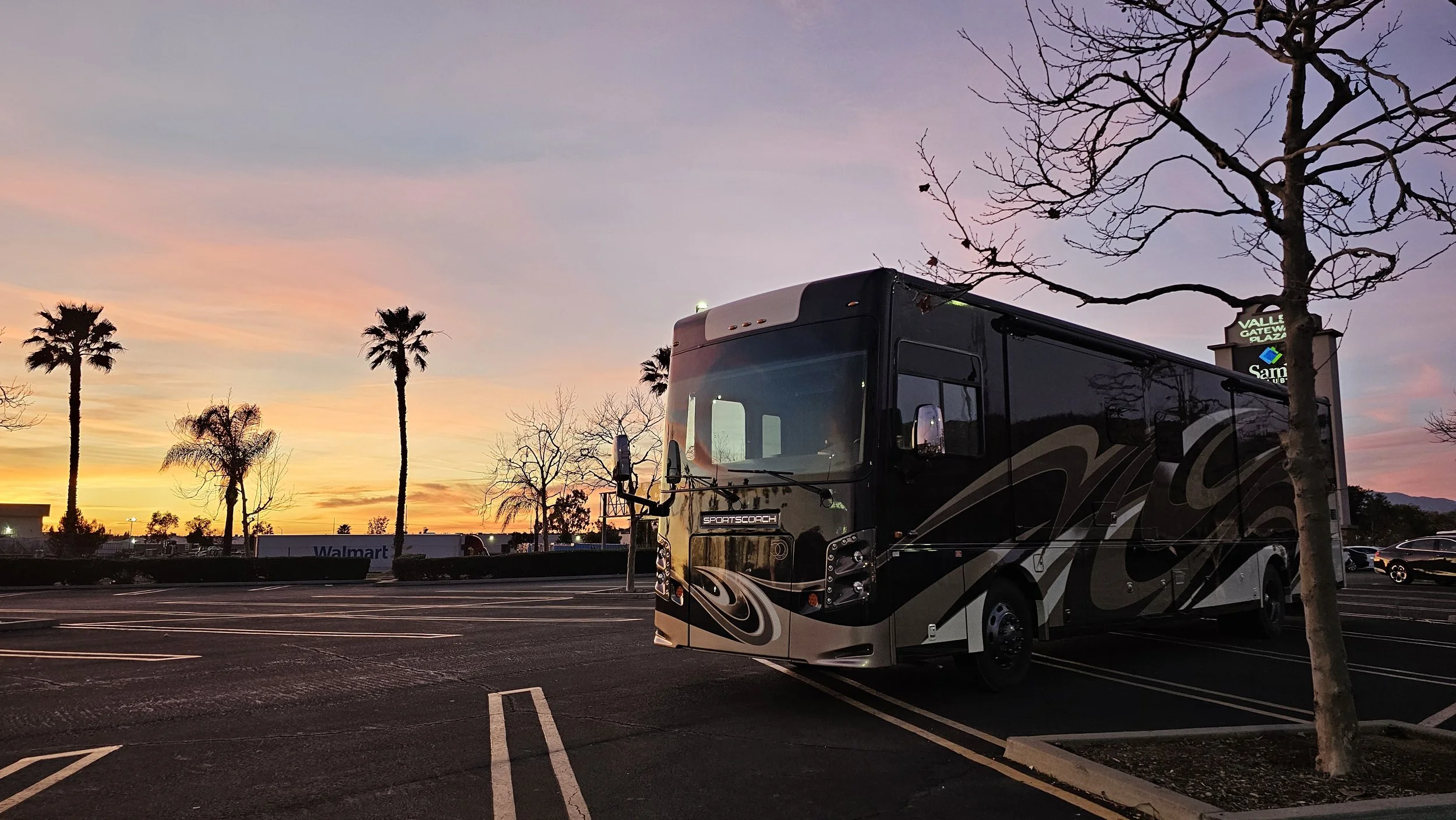 A black and gray sports coach bus parked in an empty parking lot at sunset, with silhouettes of palm trees and a leafless tree, and a Walmart store sign visible in the background.