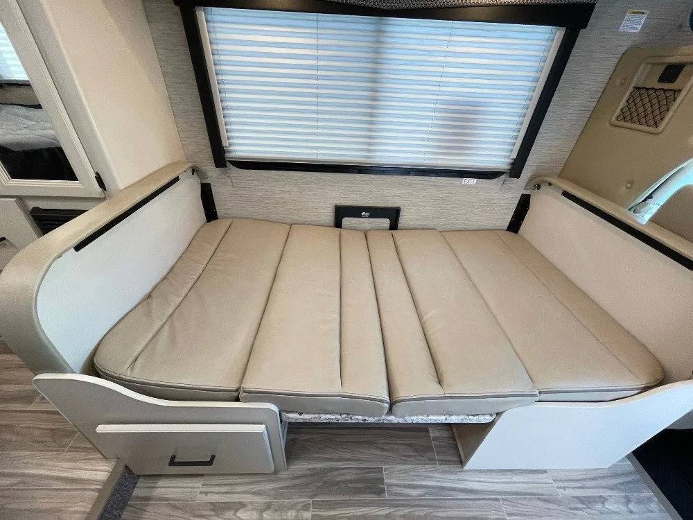 Light beige cushioned RV sofa bed with storage drawer underneath, window with closed blinds behind, and part of an interior wall and control panel visible.