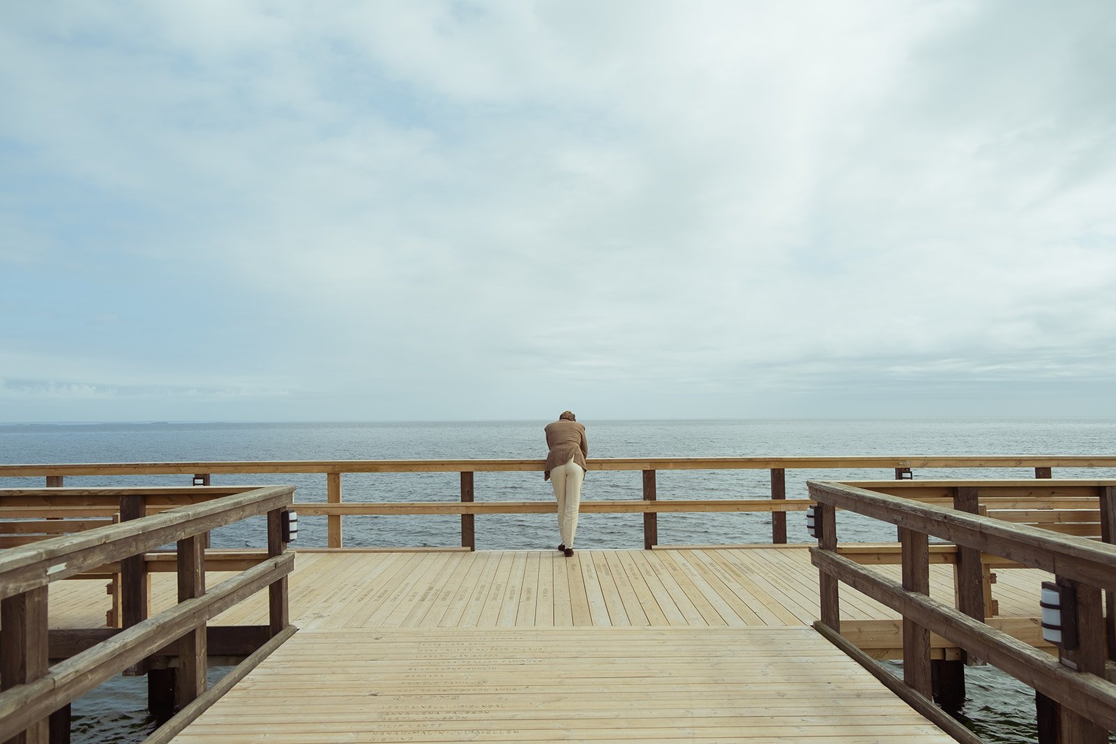 Person standing on a wooden pier looking at the ocean under a cloudy sky.