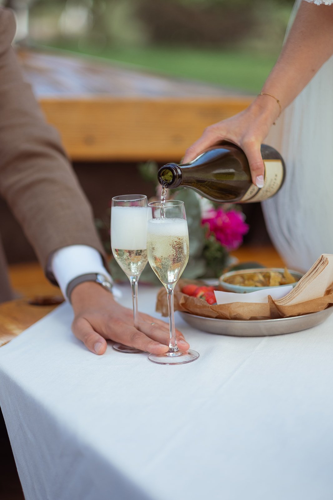 A person pouring champagne into glasses on a table with a plate of food and a white tablecloth.