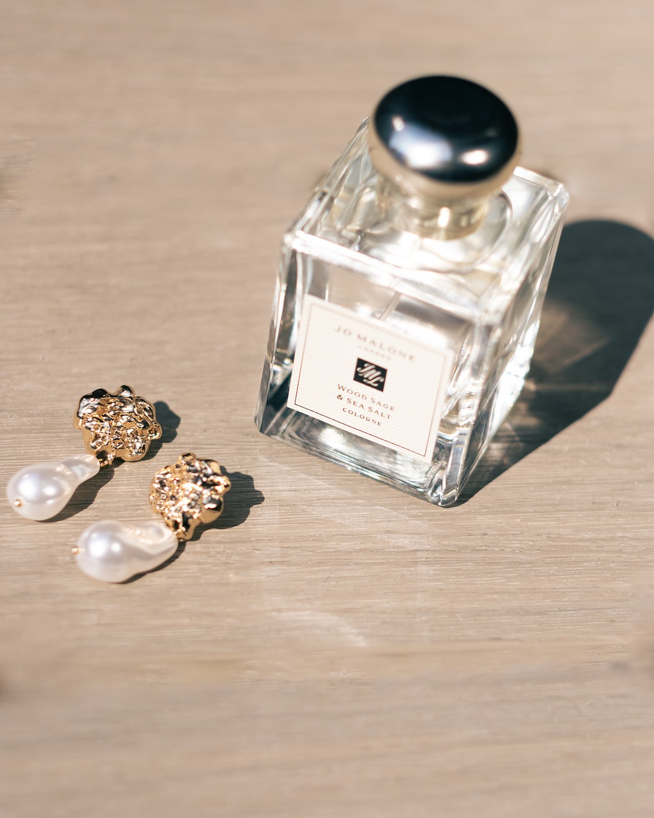 Bottle of Jo Malone Wood Sage & Sea Salt cologne next to a pair of gold and pearl earrings on a wooden surface.