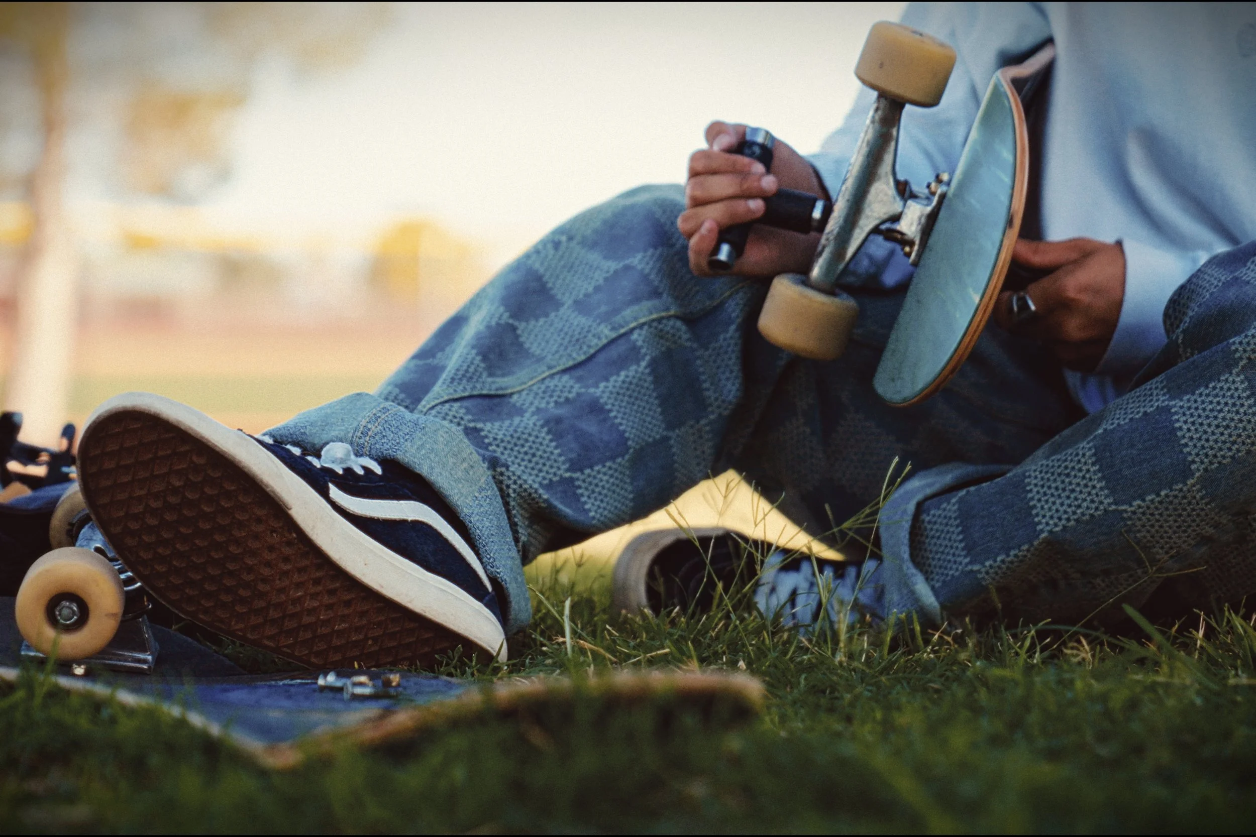Person sitting in grass holding a skateboard with a wrench, wearing patterned pants and sneakers.