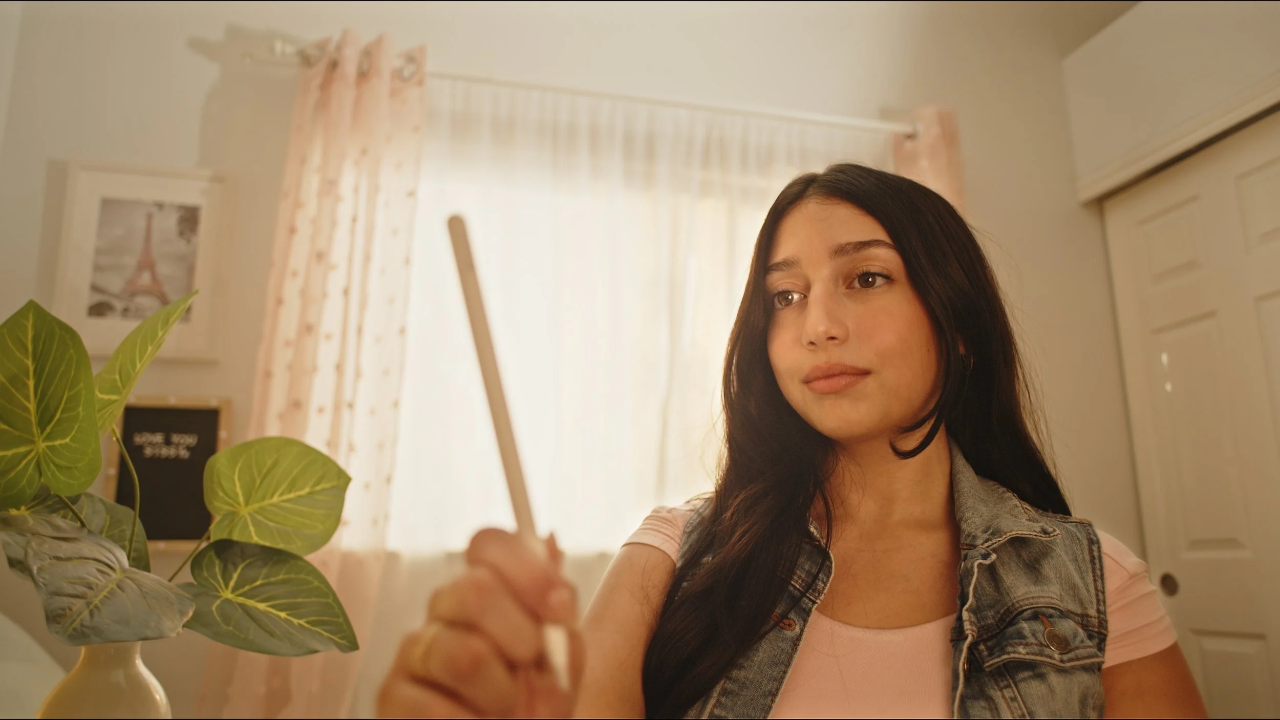 A woman with long dark hair holds a phone, taking a selfie in a warmly lit room. Behind her are pink curtains, a potted plant, and a framed picture of the Eiffel Tower.