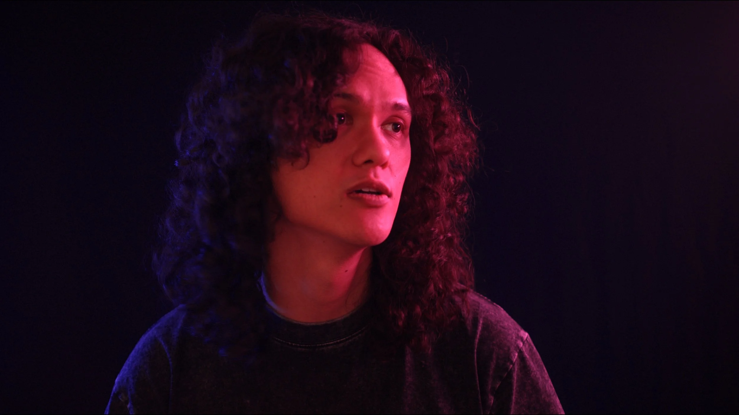 Person with long curly hair in dramatic lighting, wearing a dark shirt.