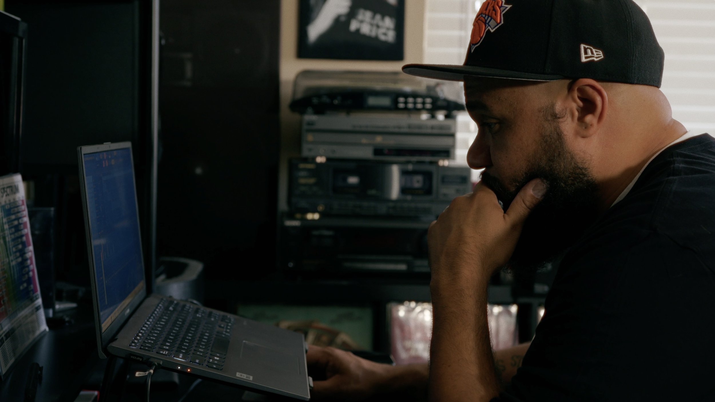 Person sitting at a computer, wearing a black cap, with audio equipment in the background.