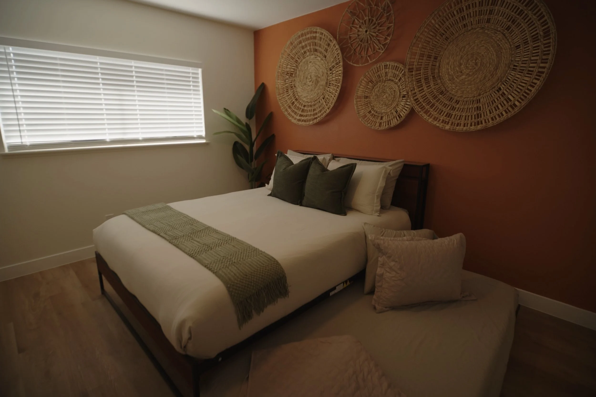 Cozy bedroom with orange accent wall, large bed with green and brown pillows, woven wall decor, and a plant by the window with blinds. Real Estate videography.