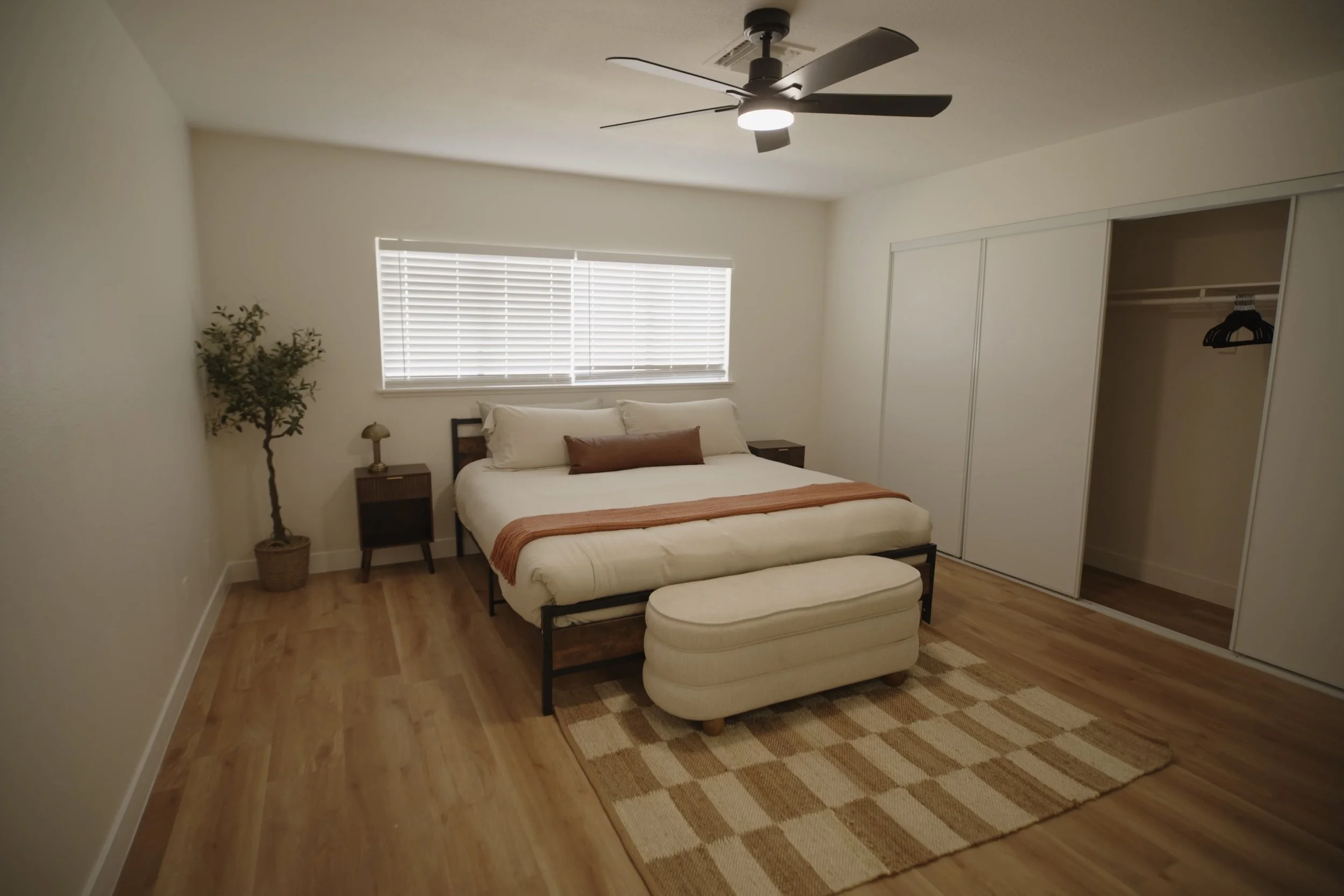 Minimalist bedroom with double bed, white and brown bedding, ceiling fan, potted plant, wooden nightstands, and an open closet. Real Estate videography.