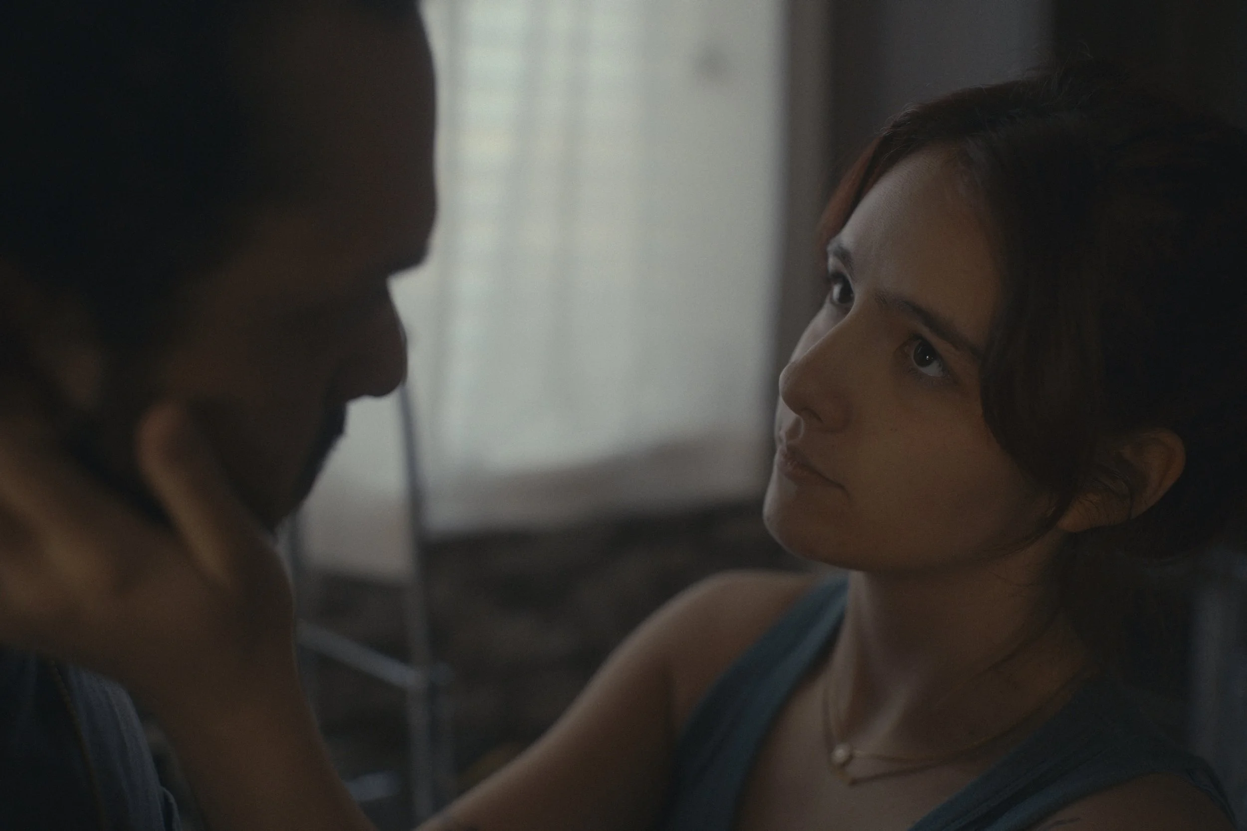 A woman with dark hair and a blue tank top looking intently at a man whose face is partially visible. They appear to be in a close, intimate moment indoors with soft lighting.