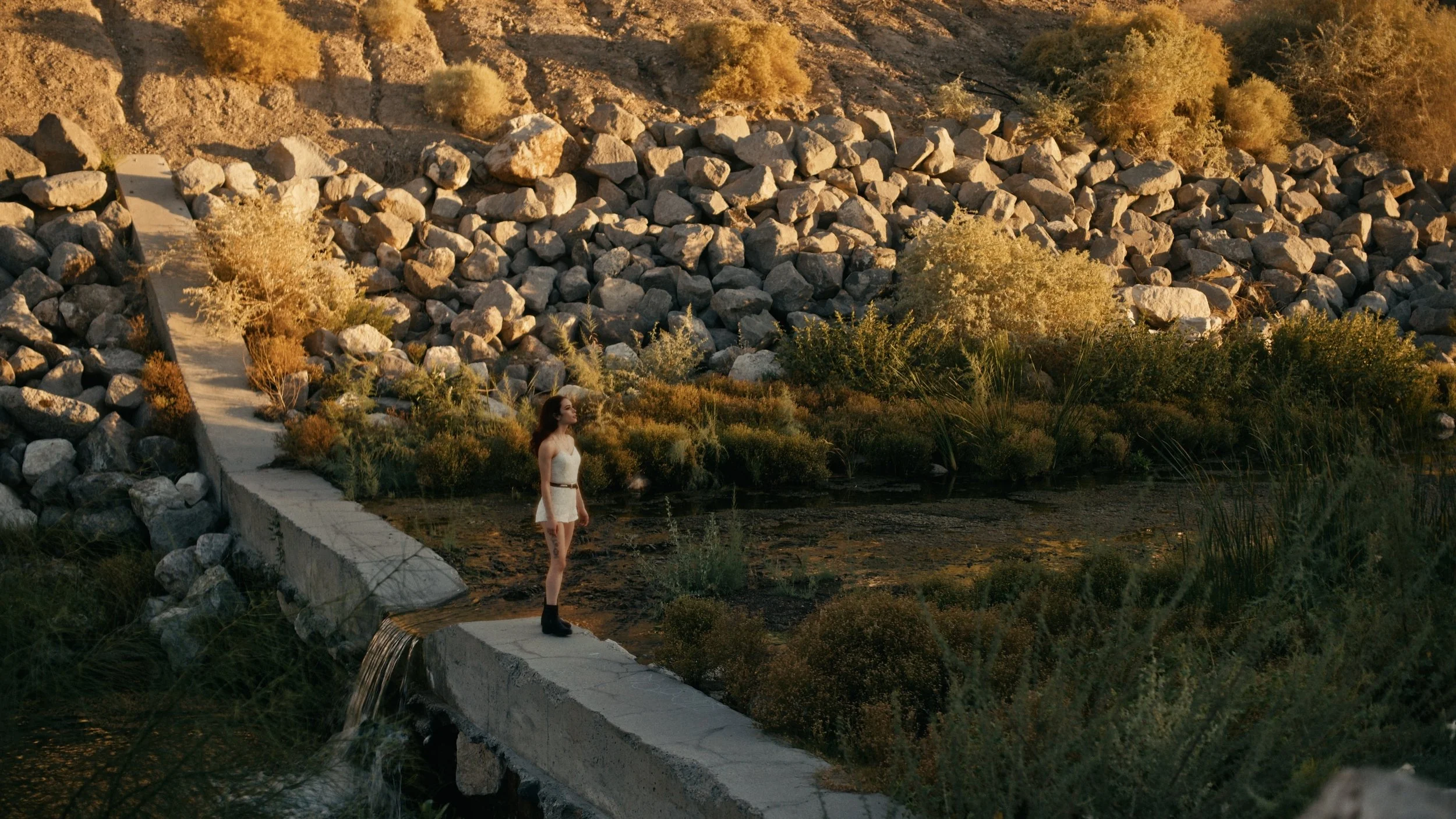 Person standing on a concrete path near a small stream, surrounded by rocks and vegetation in a dry, hilly landscape.
