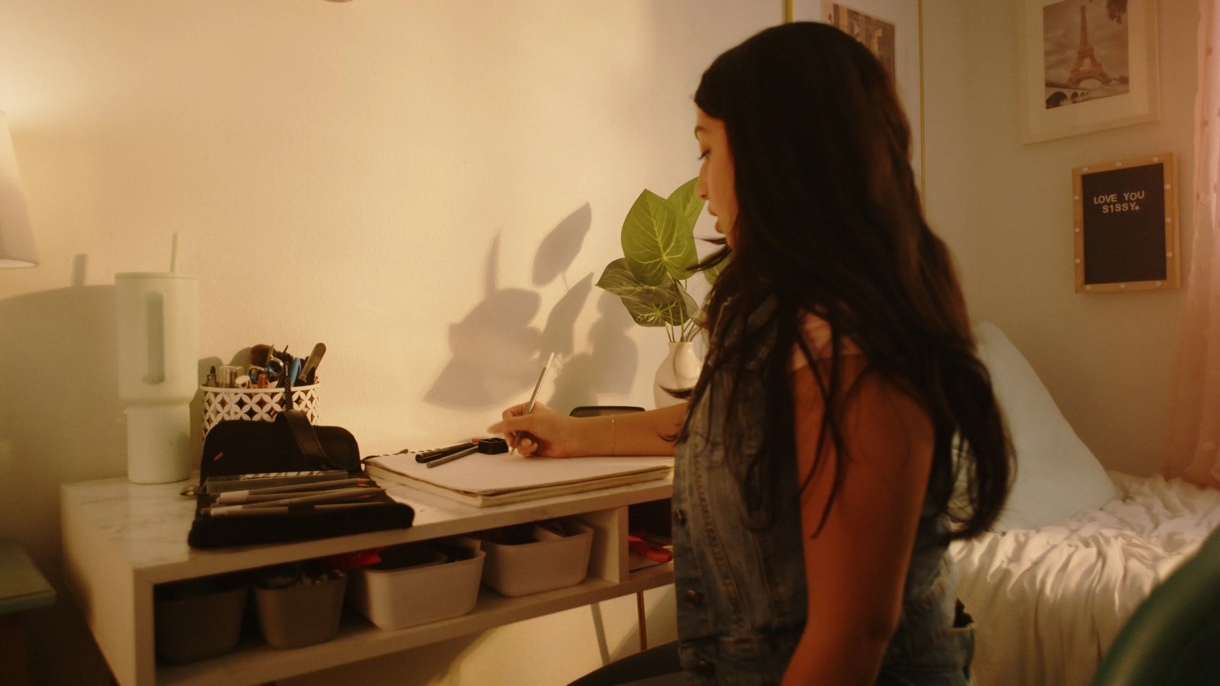 Person writing at a desk with a plant, organized stationery, and decor in a cozy room.