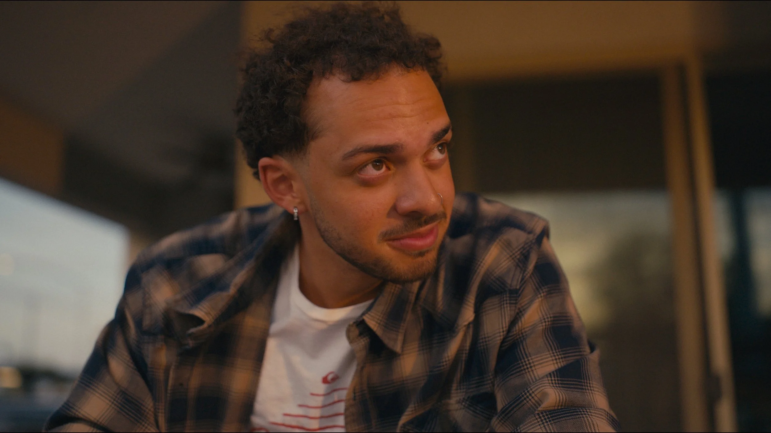 A young man with curly hair, earrings, and a nose ring looking to the side, wearing a plaid shirt over a white T-shirt, sitting indoors.