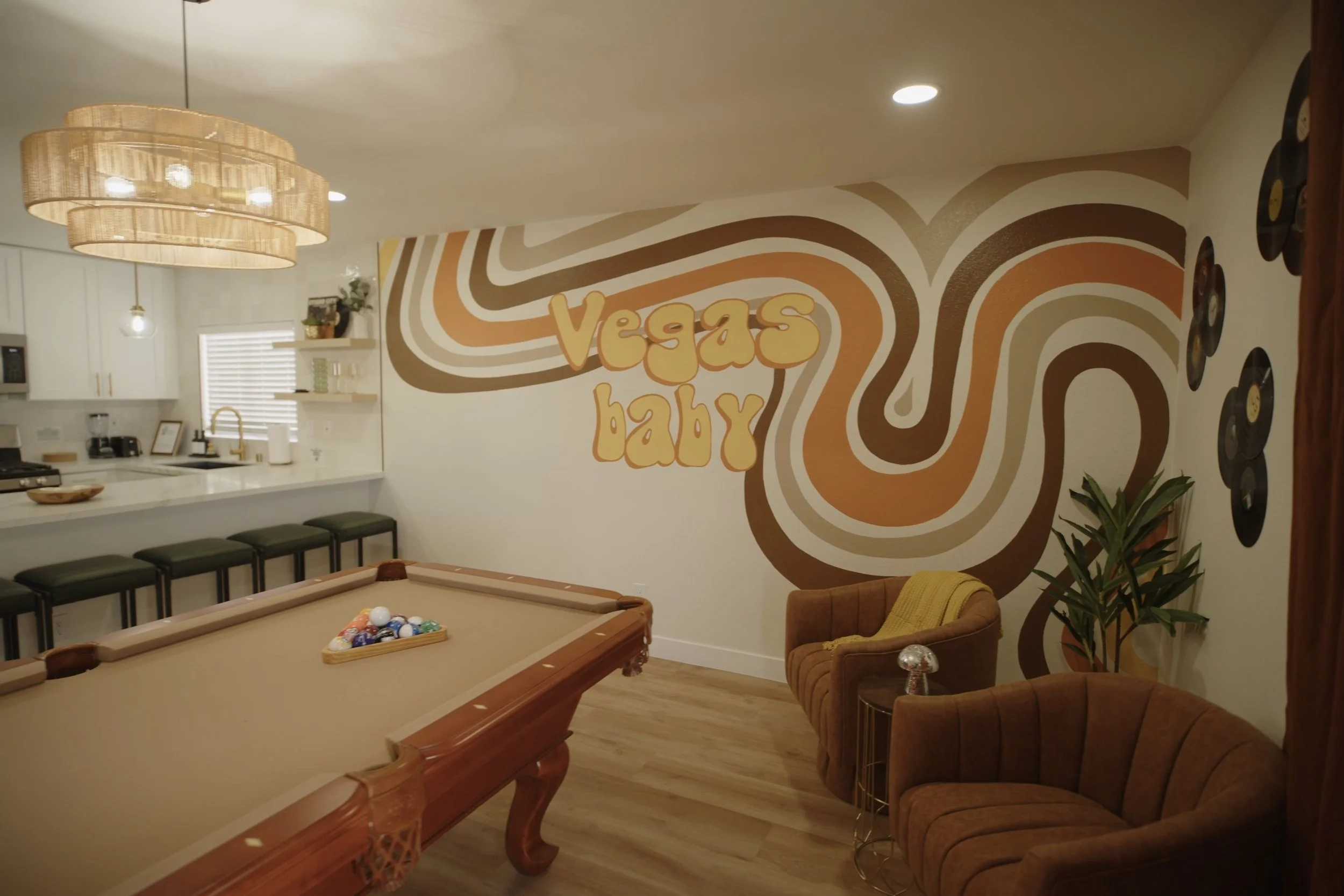 Retro-themed room with wall art that says 'Vegas baby', a pool table, brown chairs, vinyl records as decor, and a wicker chandelier. Real Estate videography.