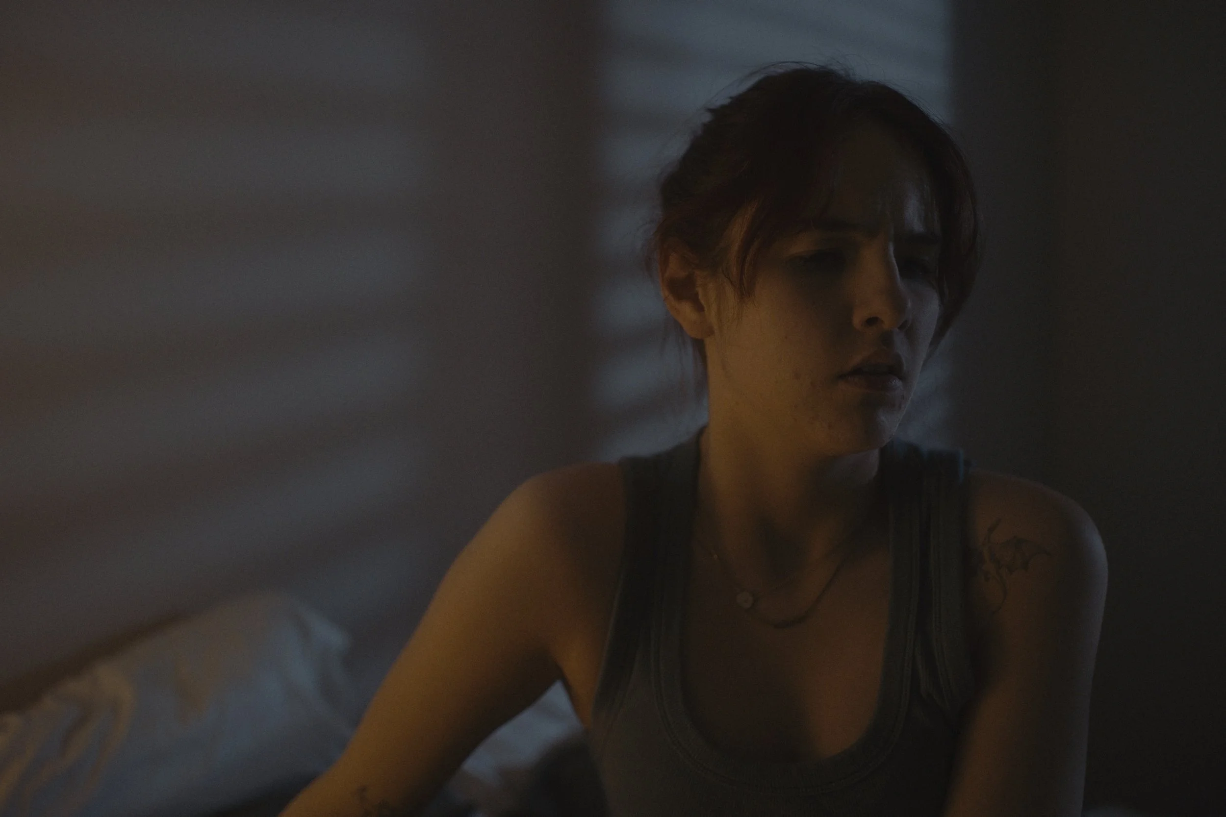 A woman with short hair and tattoos, wearing a tank top, sits in a dimly lit room with a serious or contemplative expression.