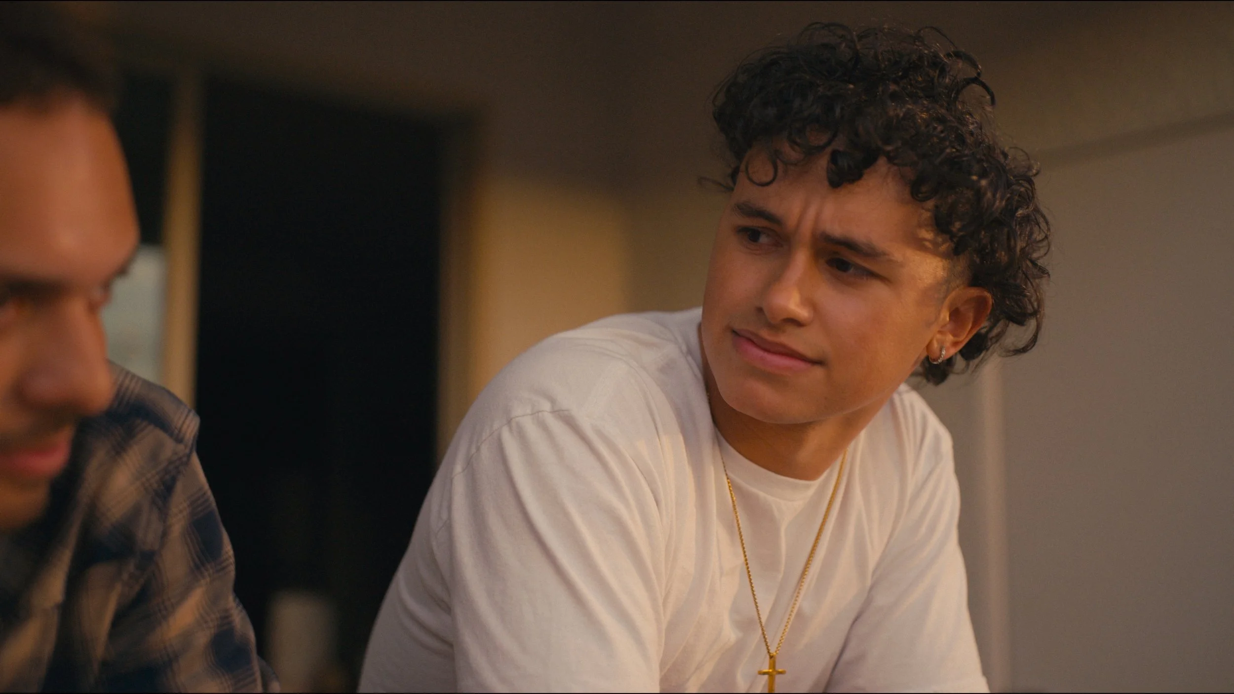 A young man with curly hair wearing a white t-shirt and gold cross necklace looking at another person, partially out of frame, in an indoor setting with warm lighting.