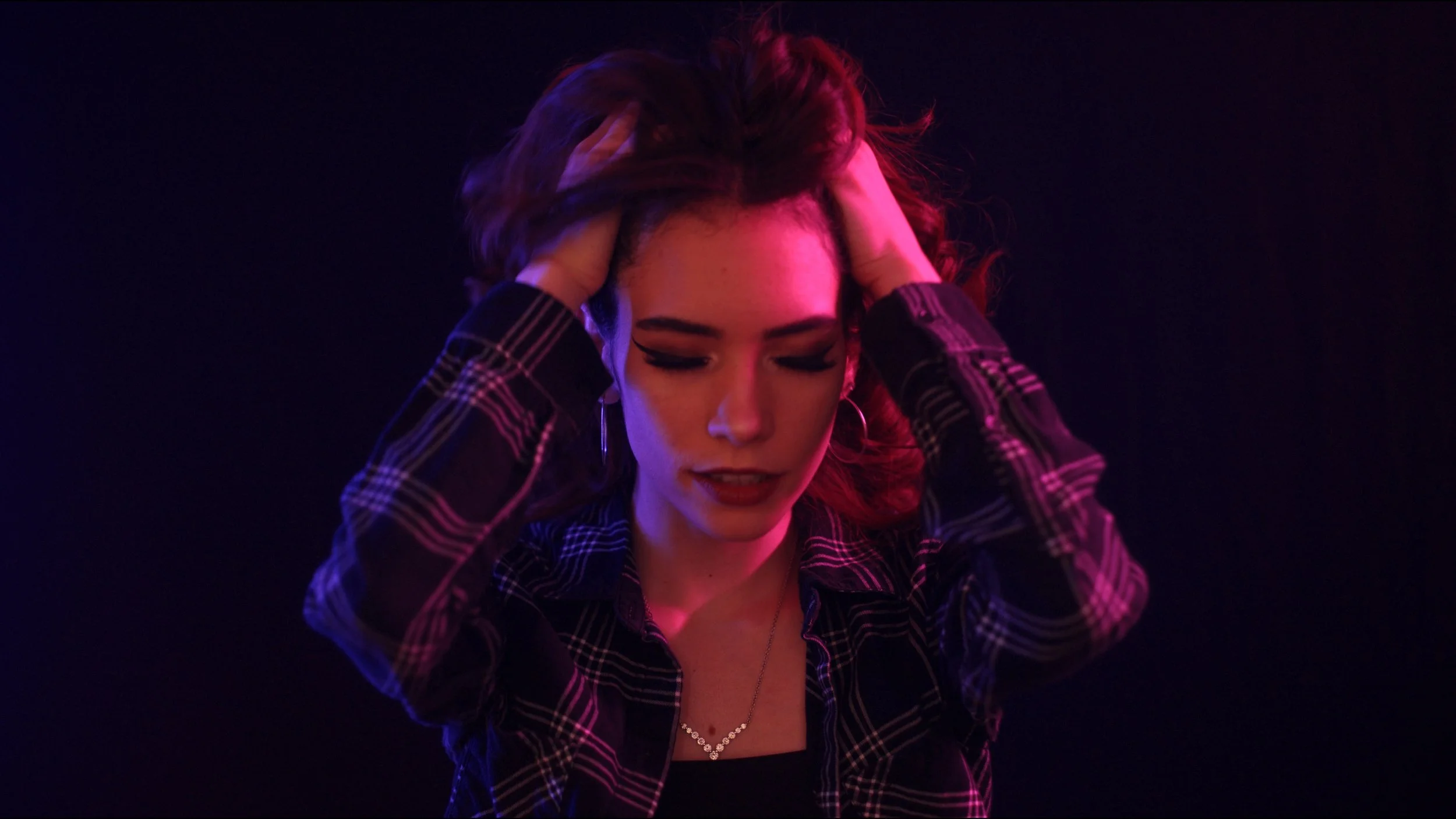 Person with long hair and plaid shirt, eyes closed, hands in hair, under pink and purple lighting