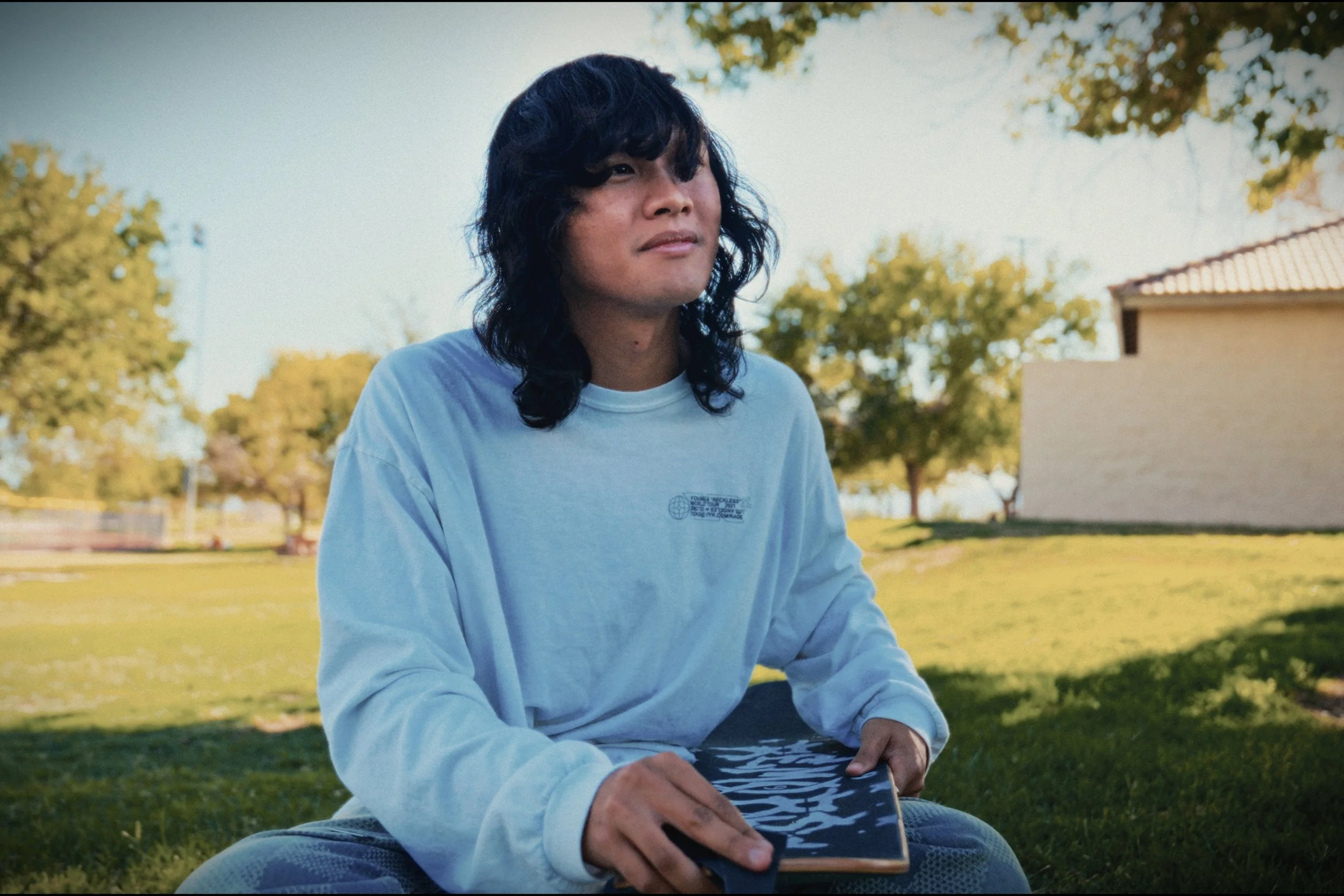 Person with long hair in a white shirt holding a skateboard, seated in a grassy park area.