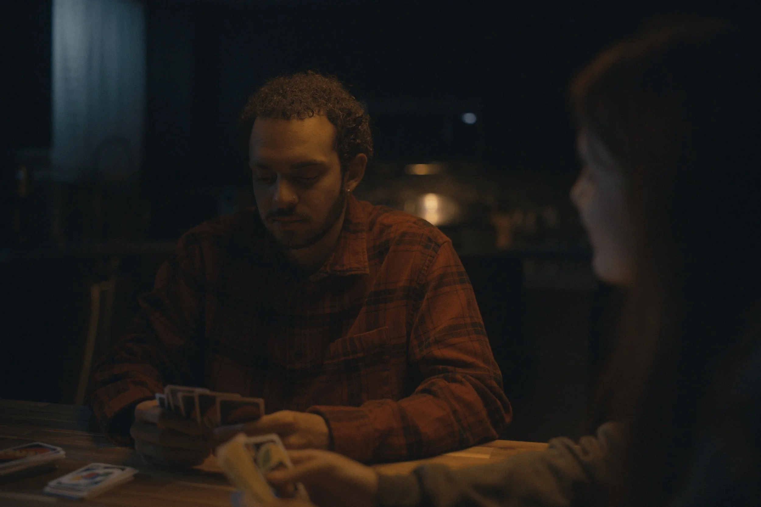 Two people playing cards in a dimly lit room, one man and one woman, with the man wearing a plaid shirt.