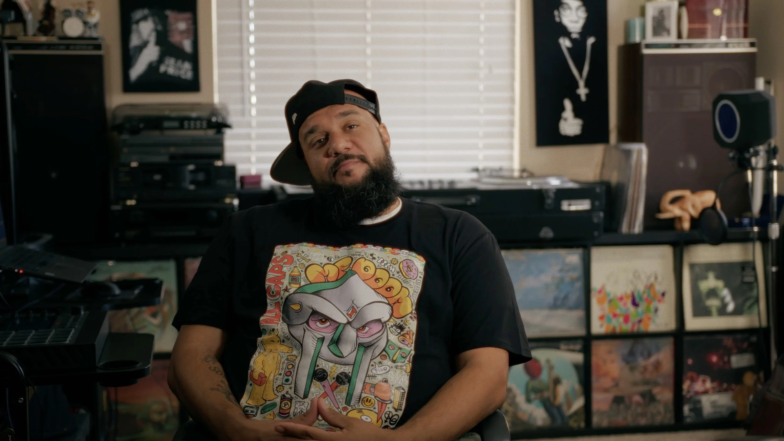 Man with beard and baseball cap in a music-themed room, wearing a graphic t-shirt, with vinyl records and audio equipment in the background.