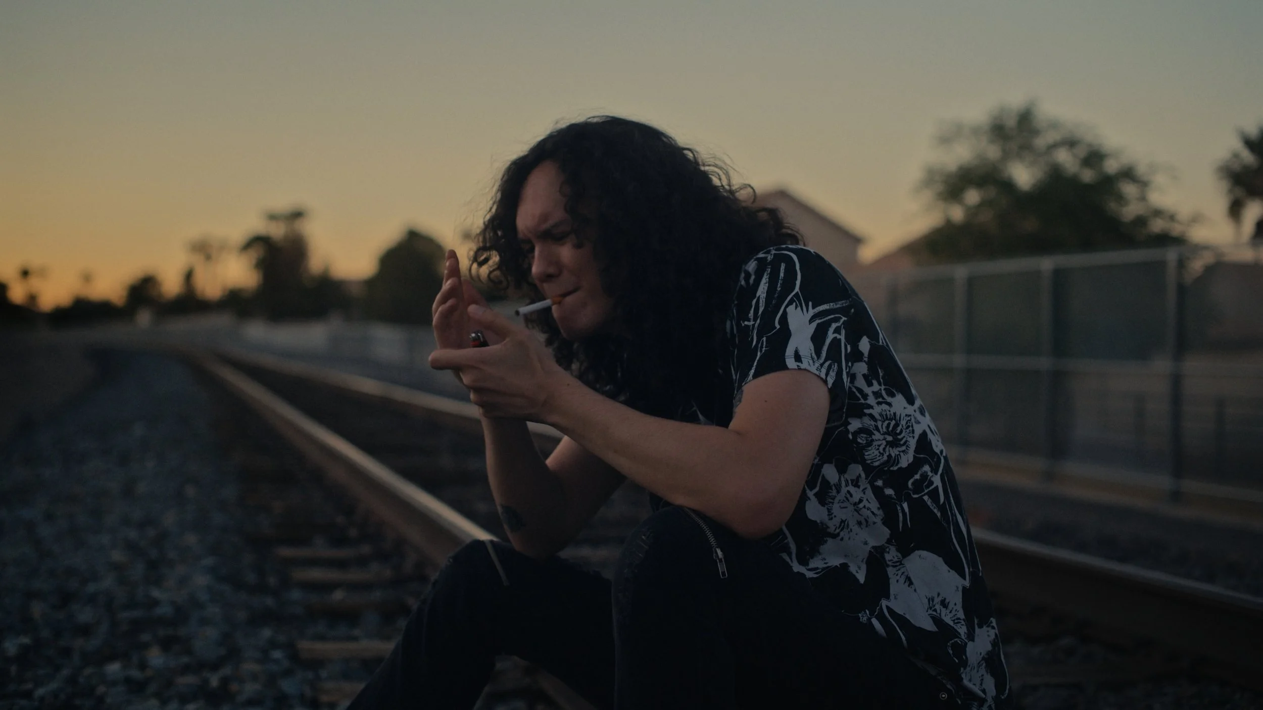 Person with long curly hair sitting on railroad tracks, attempting to light a cigarette with a lighter at sunset.