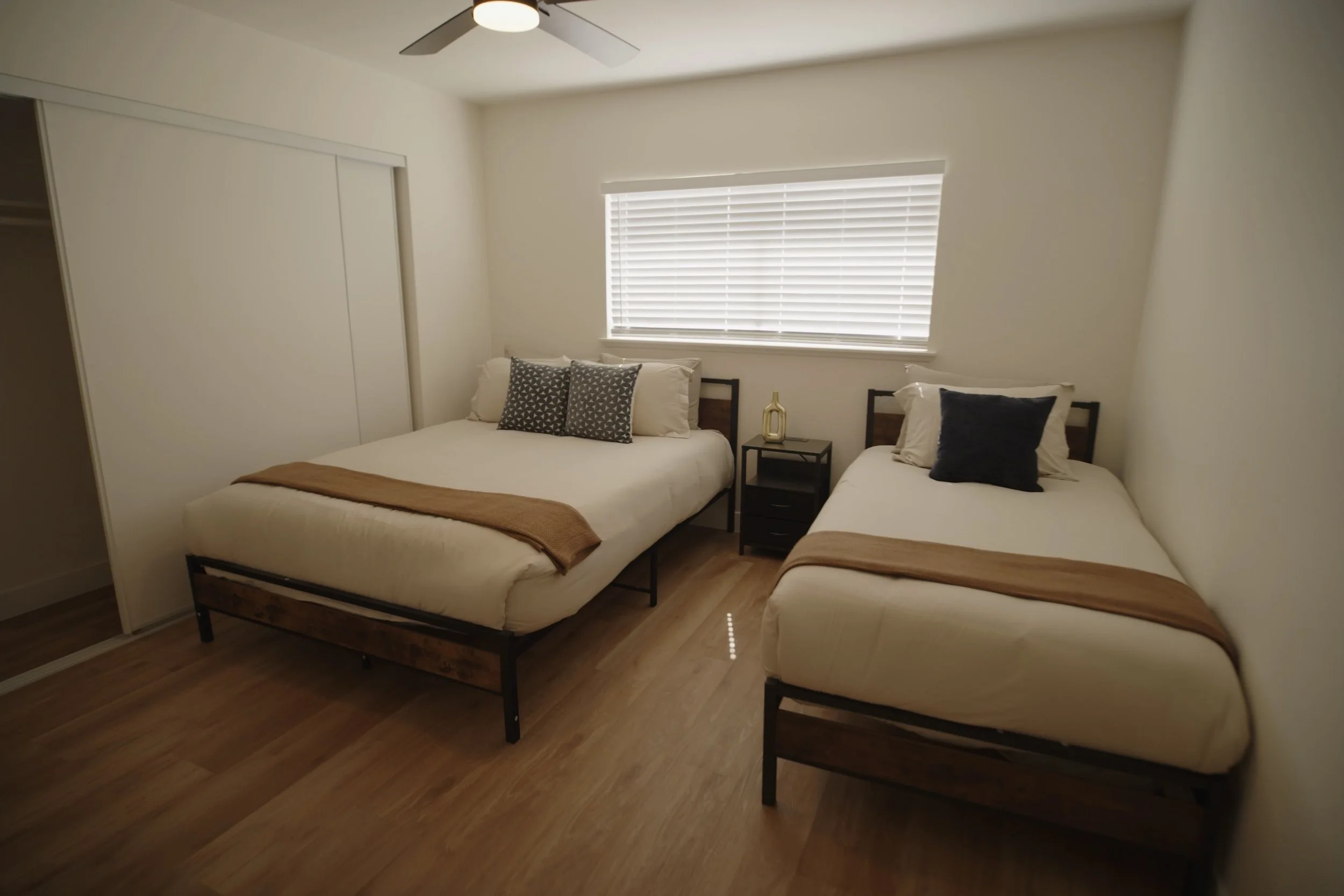 Bedroom with two beds, wooden floors, white walls, and a ceiling fan. Beds feature white linens, decorative pillows, and brown throws. A small bedside table with a decorative item is between the beds. Window with blinds lets in light from outside.
