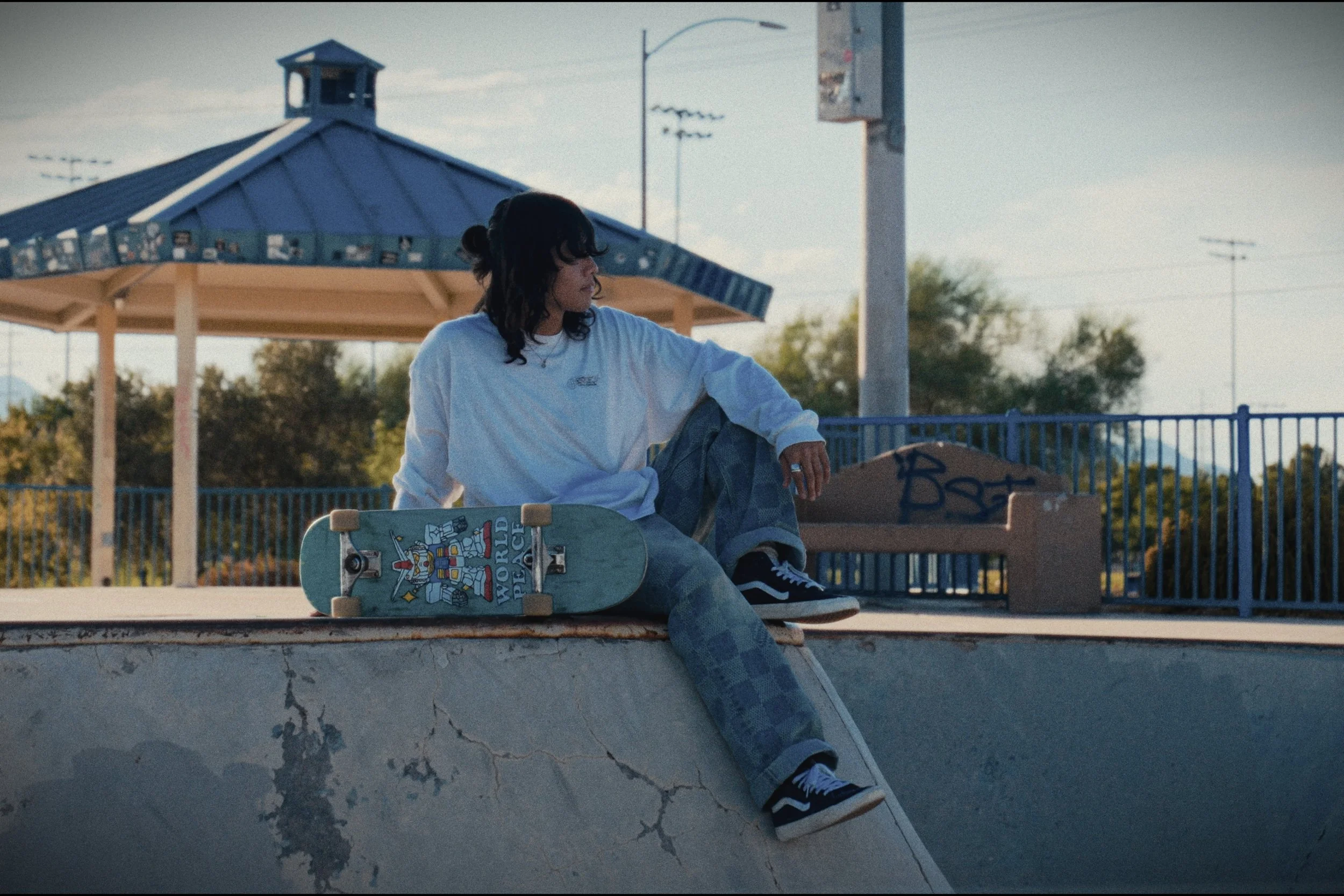 A person sitting on the edge of a skateboard ramp, with a skateboard next to them, wearing a white long-sleeve shirt and patterned pants at a skate park.