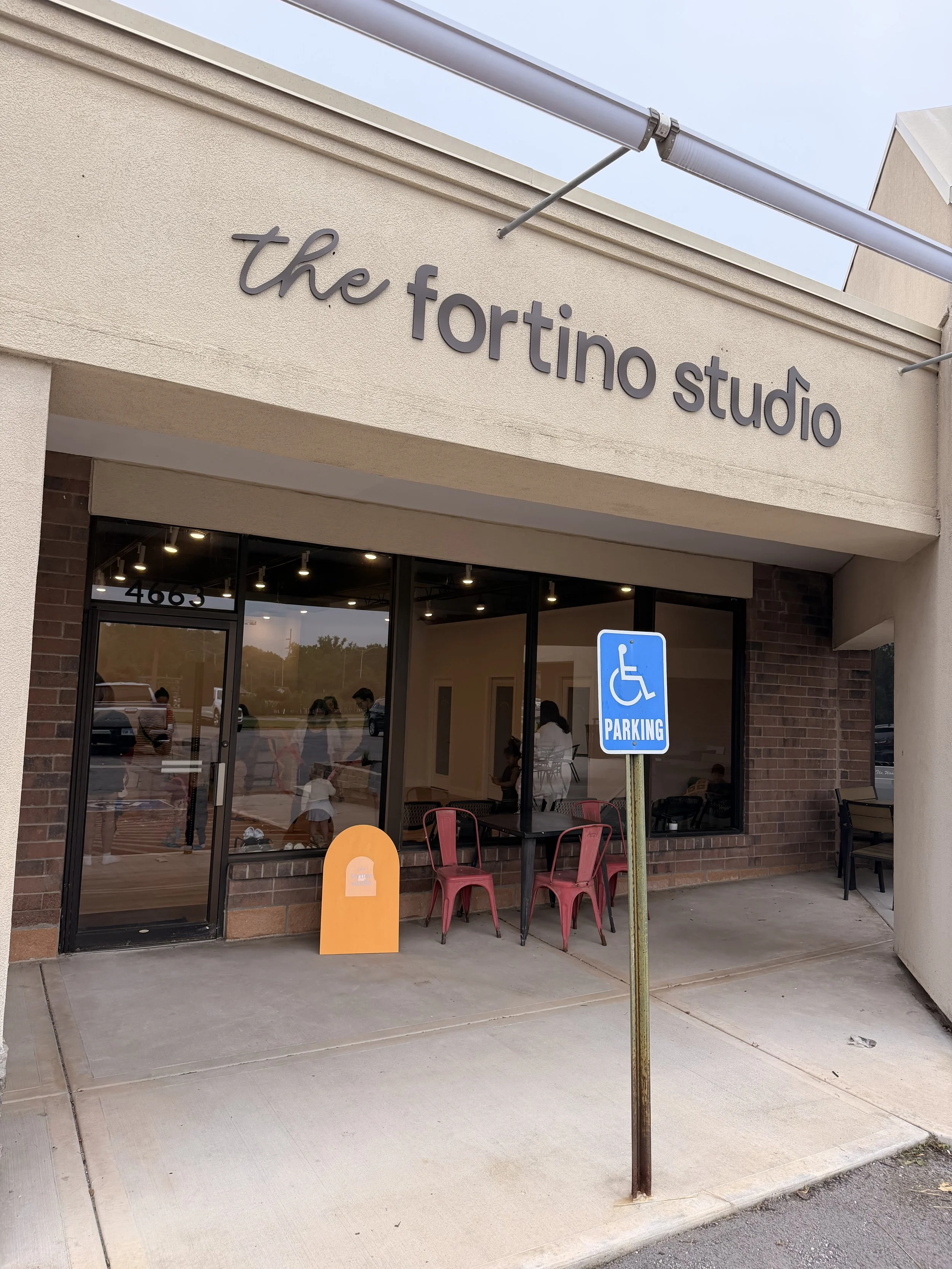 Exterior view of a bakery or cafe named 'the fortino studio', with large windows, outdoor seating with red chairs, a handicap parking sign, and a bright, clear sky.