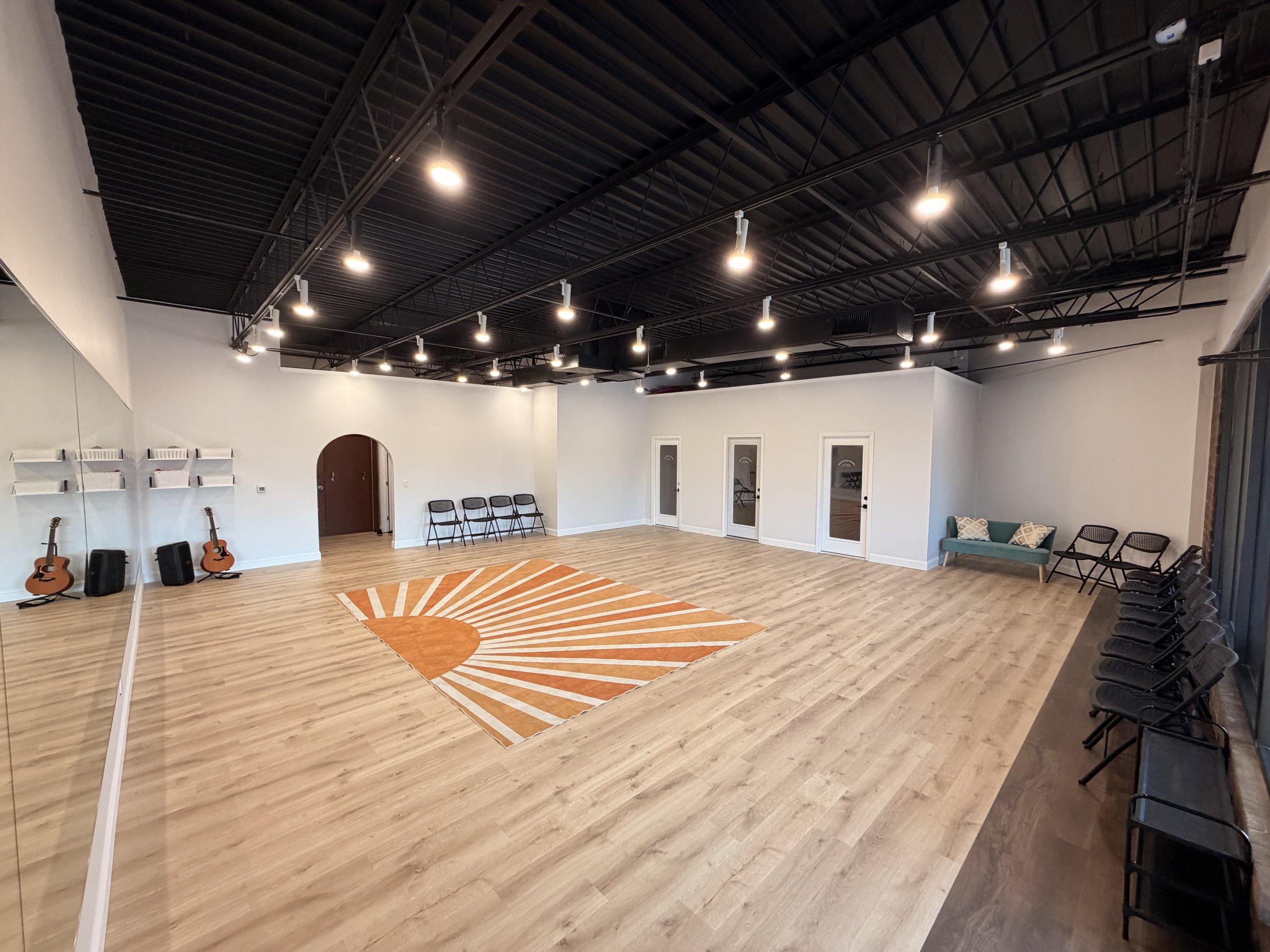 A spacious dance or yoga studio with a black ceiling and bright ceiling lights. The floor is wooden and has an orange and white circular rug. There are several chairs along the right wall, a small seating area with a green couch and pillows, and a wall mirror on the left with guitars and speakers underneath.