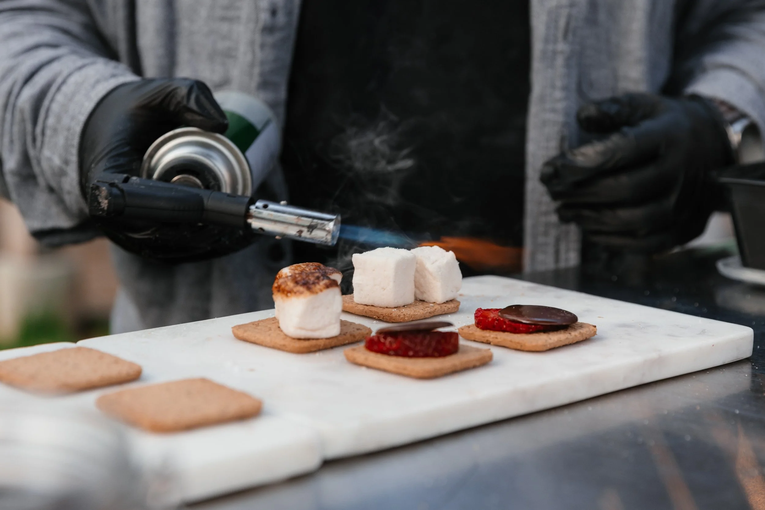 Person using a torch to caramelize marshmallows on graham crackers with chocolate, preparing s'mores.