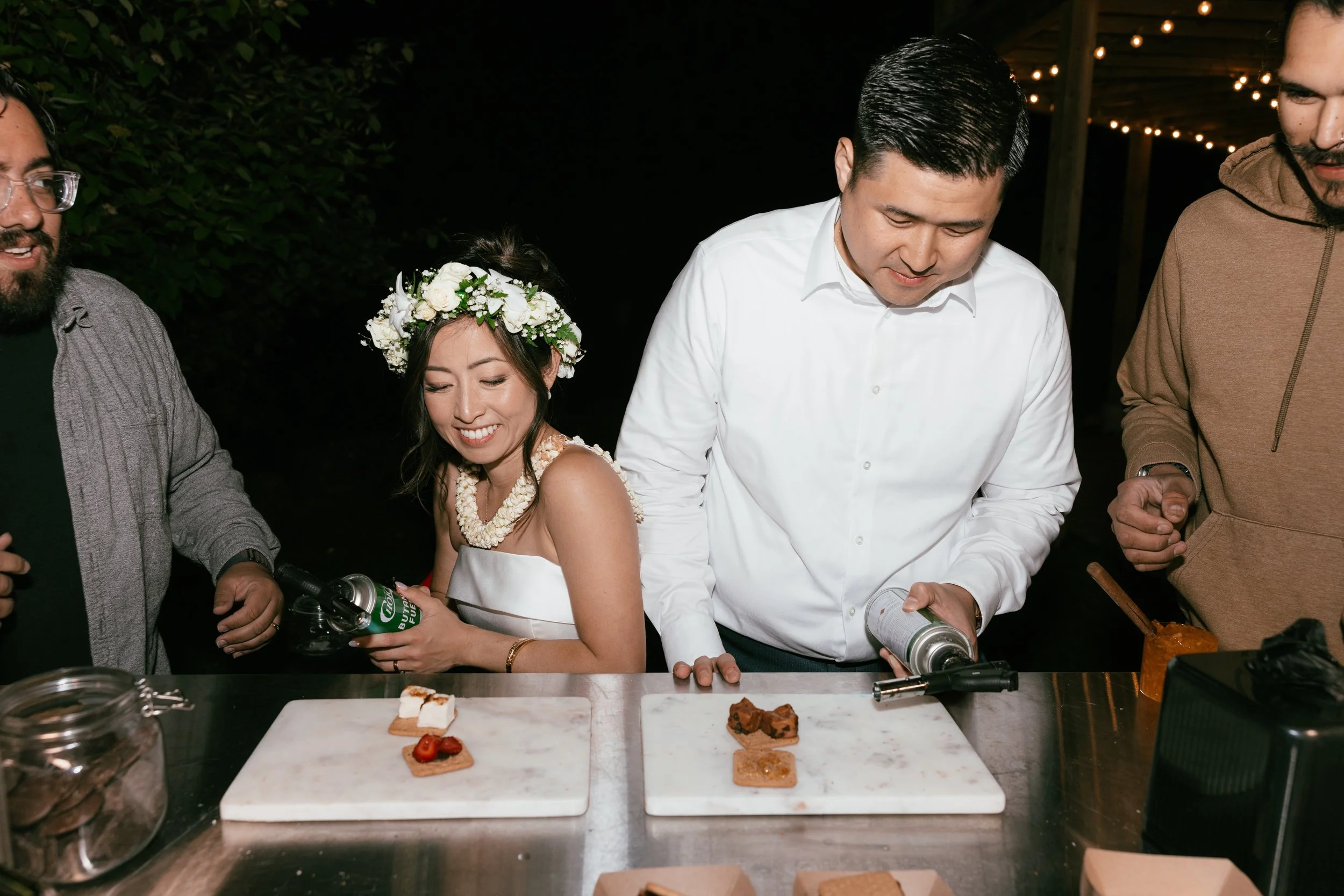 People at a celebration, woman wearing a floral crown and white dress, others in casual and formal attire, serving and preparing desserts on a table at night.