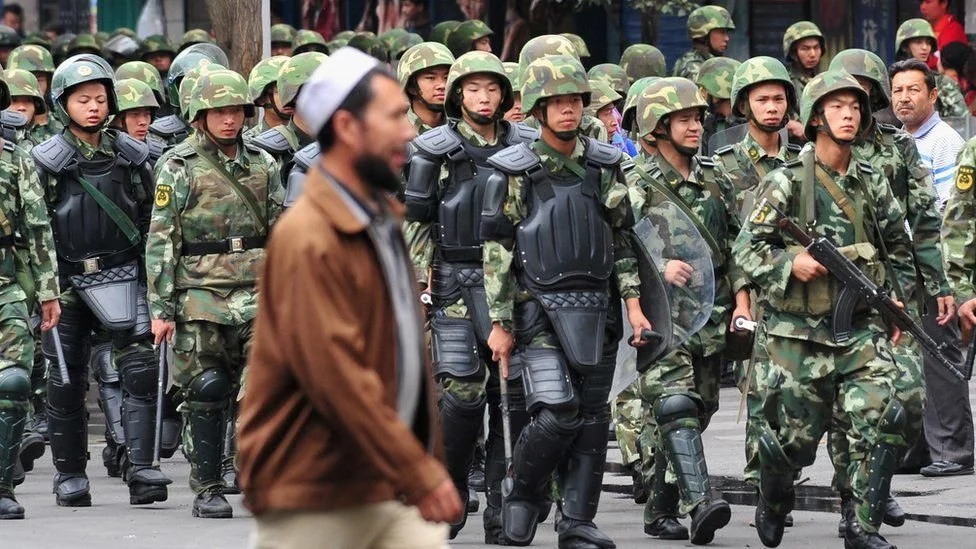 Uyghur man, blurry in foreground, walks by group of Chinese military people in uniform and tactical gear.