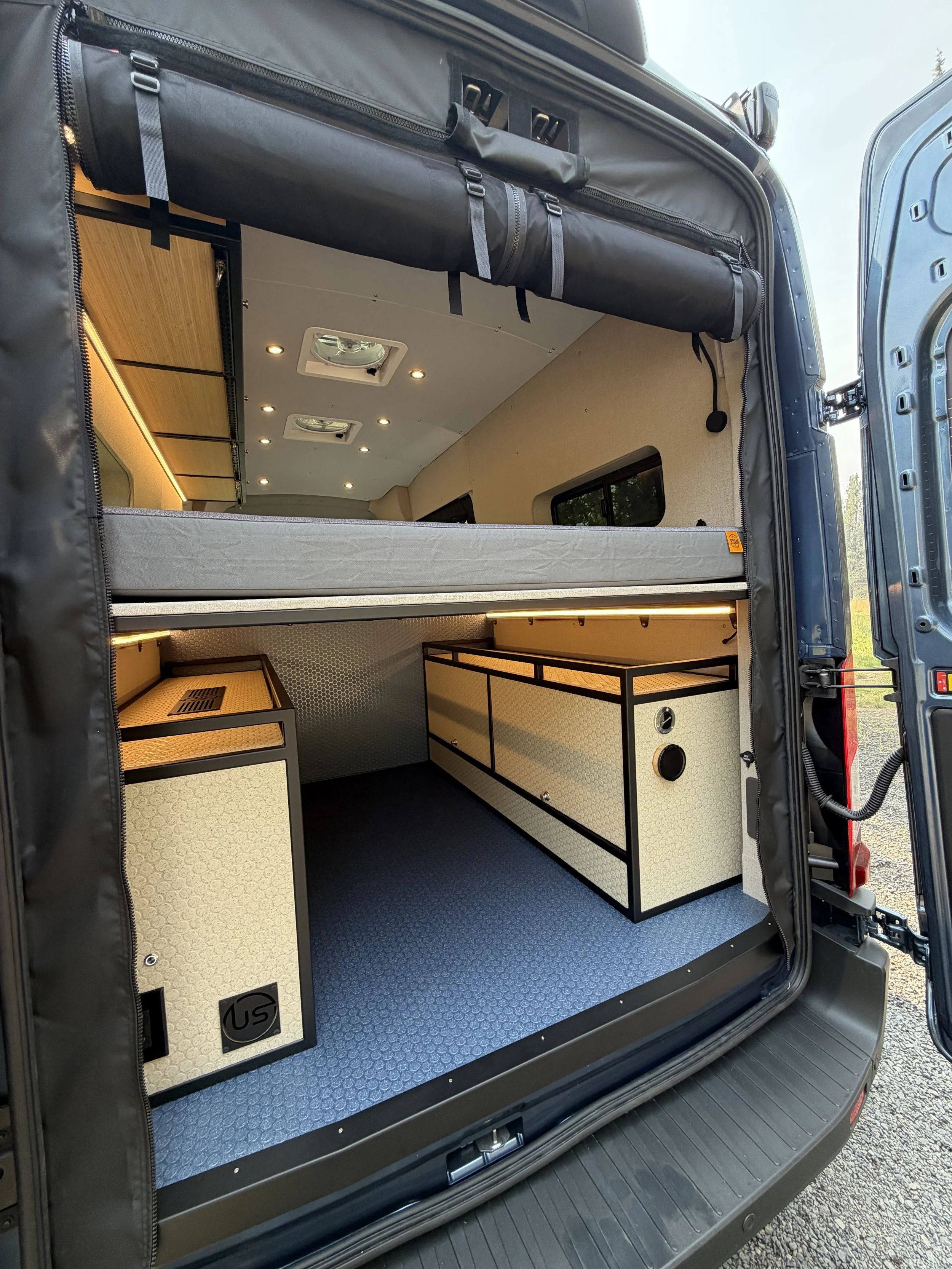 The image shows the interior of a converted van with a sleeping area, storage cabinets, and a ceiling with built-in lights and vents.