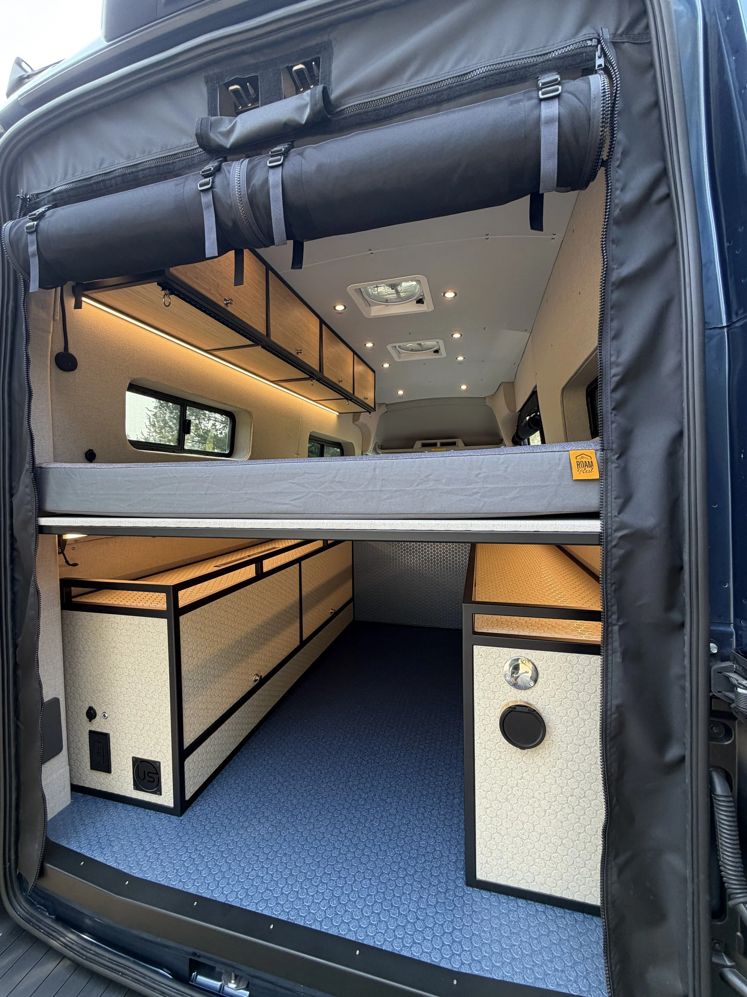 Interior of a camper van showing built-in storage cabinets, small windows, and a blue textured flooring.