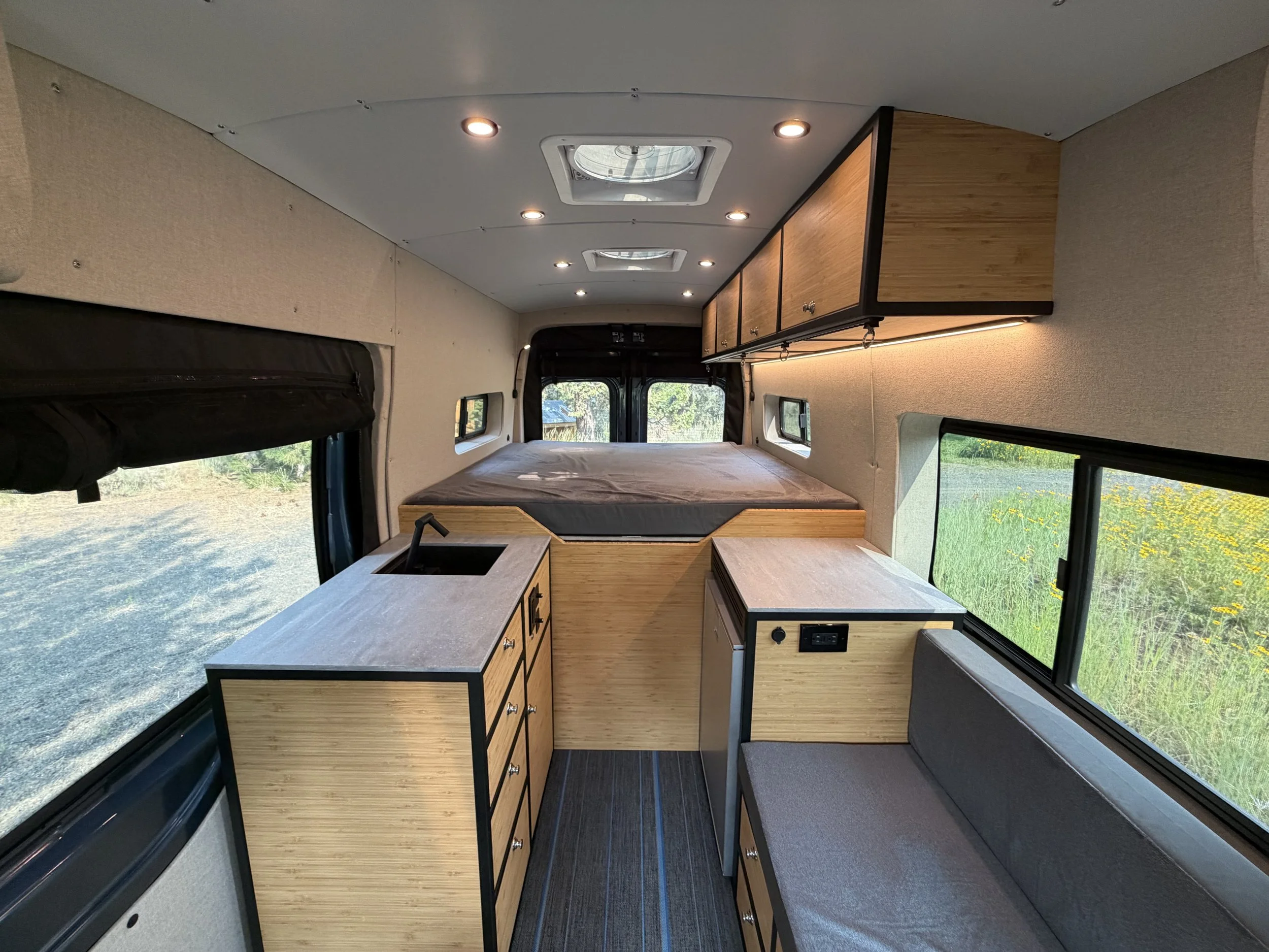 Interior of a camper van with a bed, wooden cabinets, windows, and a countertop, and skylights on the ceiling.