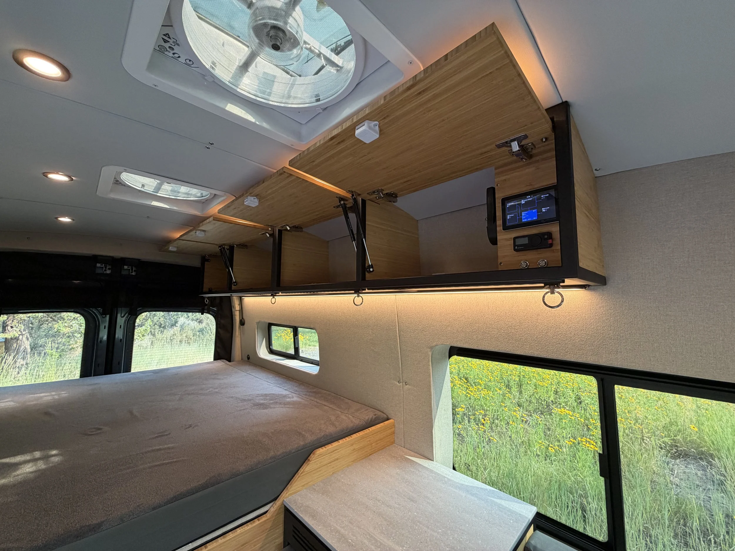 Interior view of a camper van with a wooden ceiling, a bed with gray mattress, and large windows showing a grassy field with yellow flowers outside.