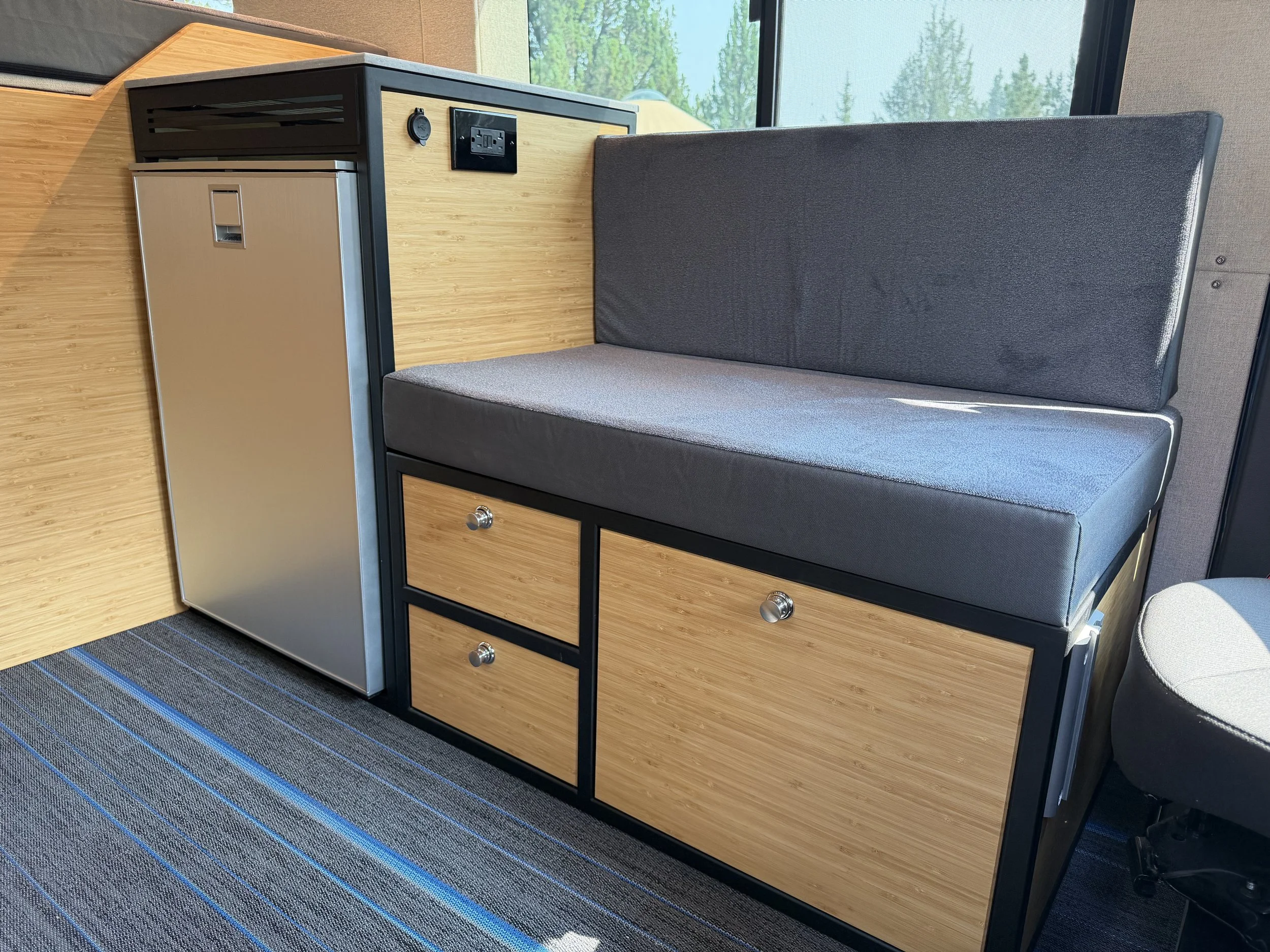 A modern workspace with a black-framed desk featuring a natural wood surface, a built-in mini fridge, and drawers with metallic handles. A gray upholstered bench is positioned beside the desk, and a large window with a screen shows green trees outside. The area has a dark striped carpet.
