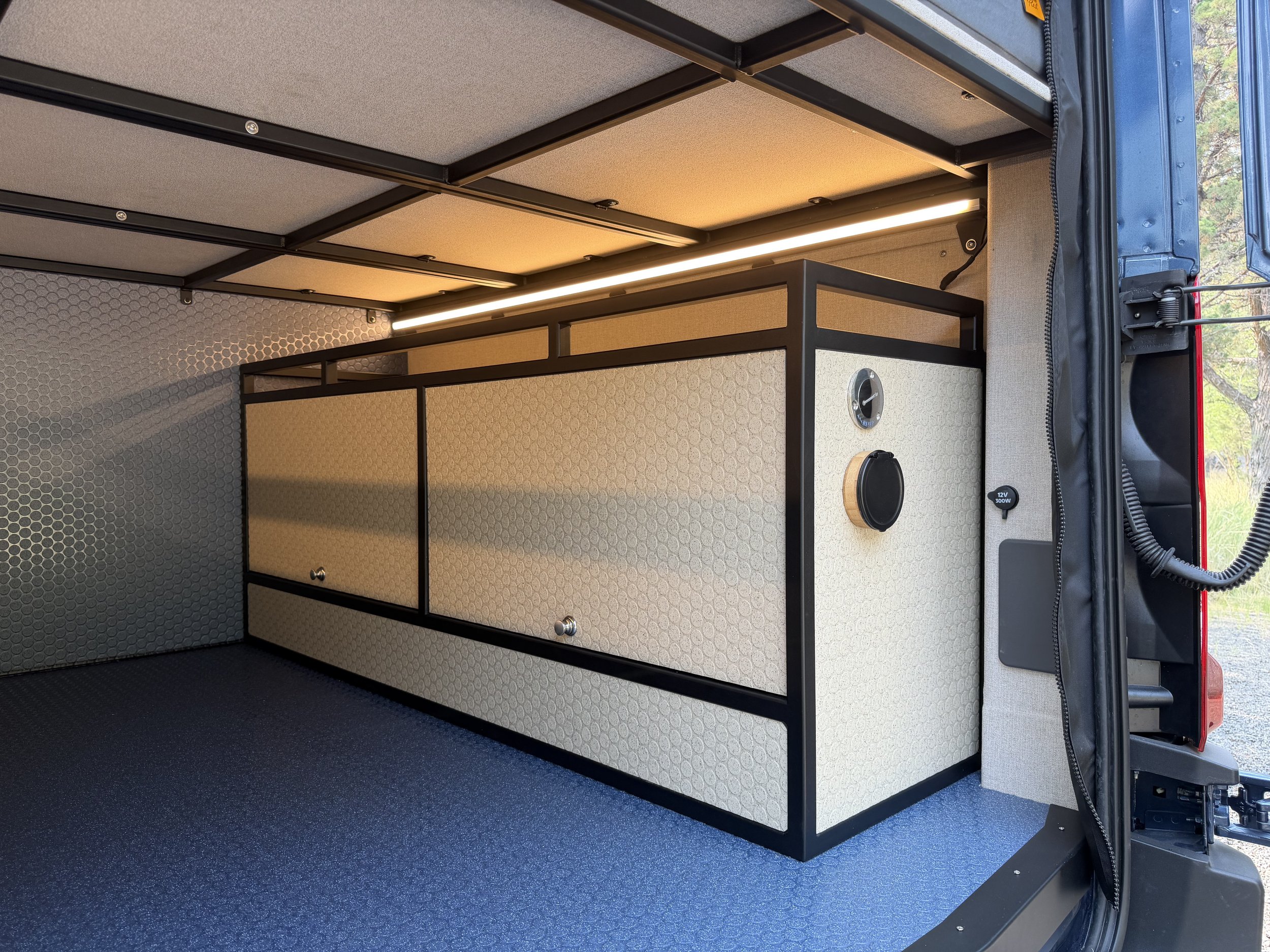 Interior of a van with a custom-built storage cabinet and a black metal frame, illuminated by a strip light. The van's side door is open, revealing trees outside.