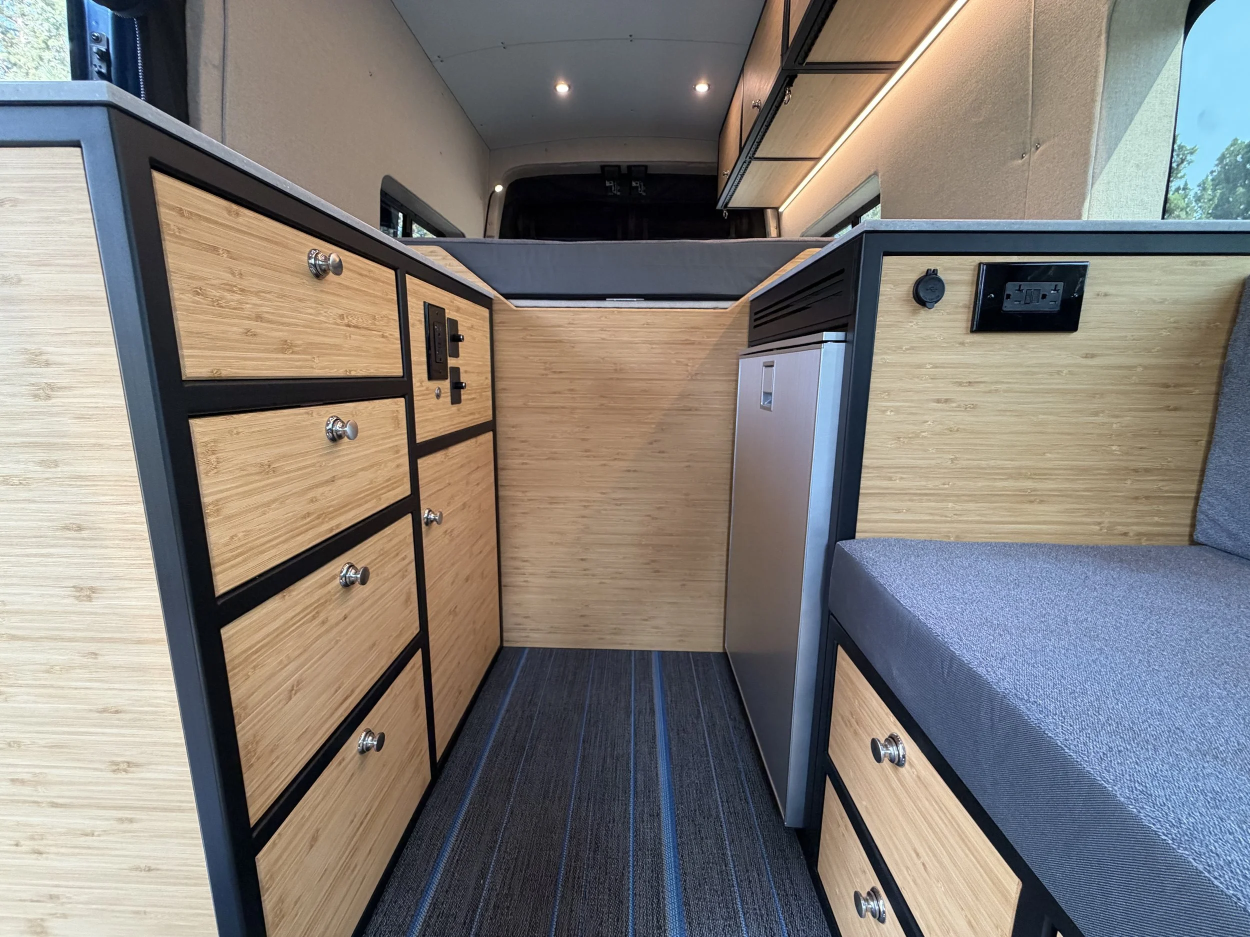 Interior view of a modern camper van with wooden cabinetry, drawers, a black countertop, and a seating area with a cushion, with windows showing trees outside.