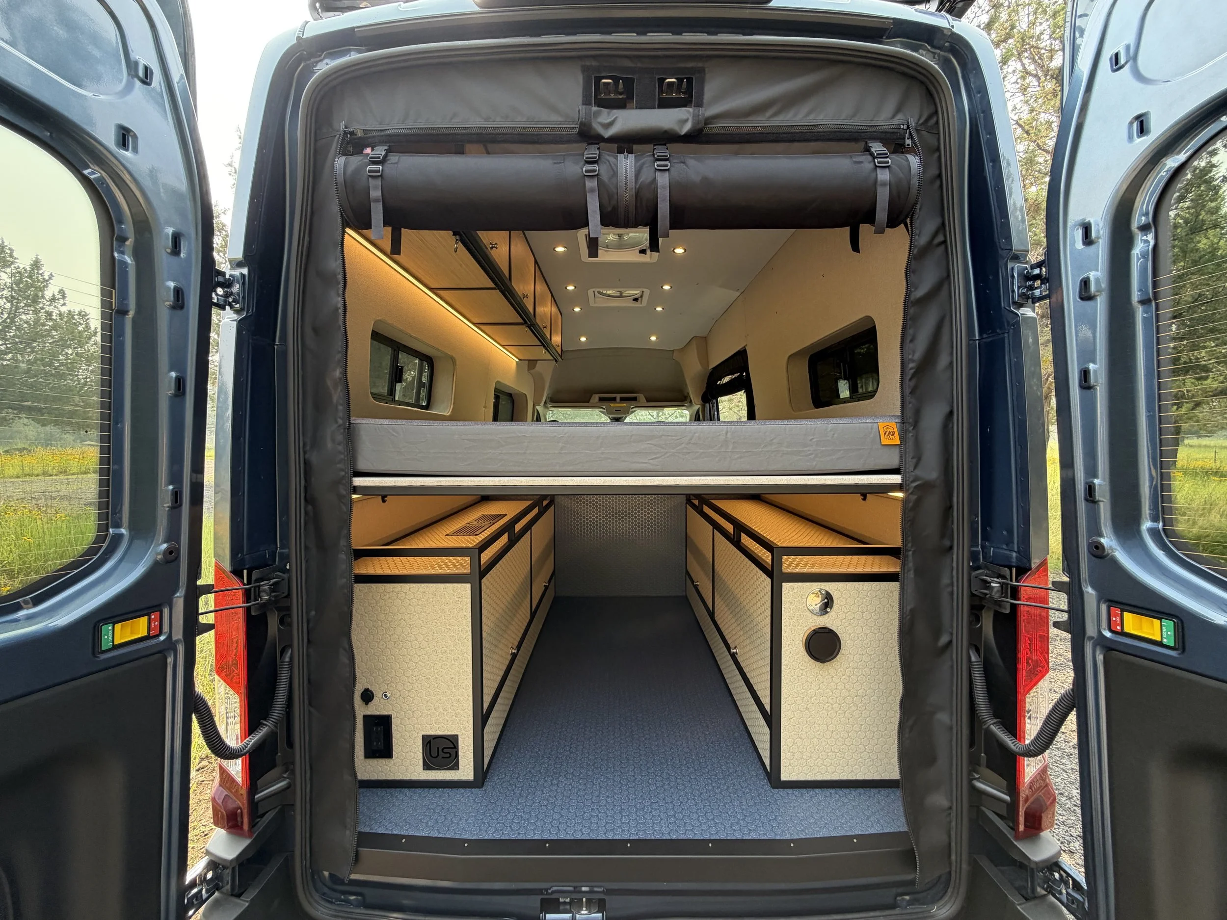 The interior of a camper van with organized storage, wood cabinetry, and a sleeping area, viewed from the open rear doors.