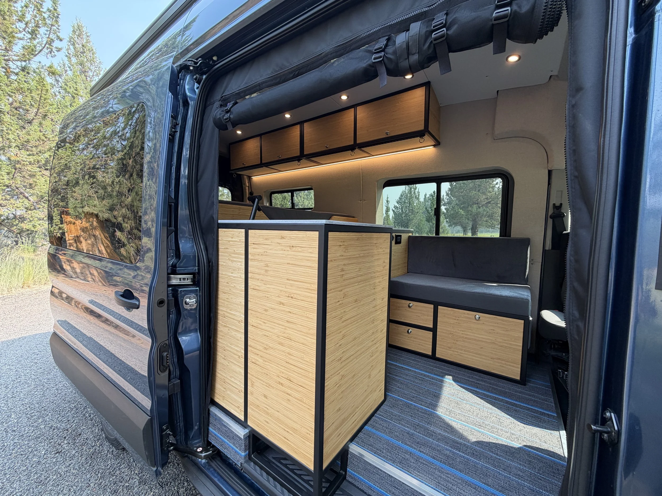 The interior of a campervan shows wooden cabinets, a small workspace, and seating, with windows revealing a forested outdoor scene.