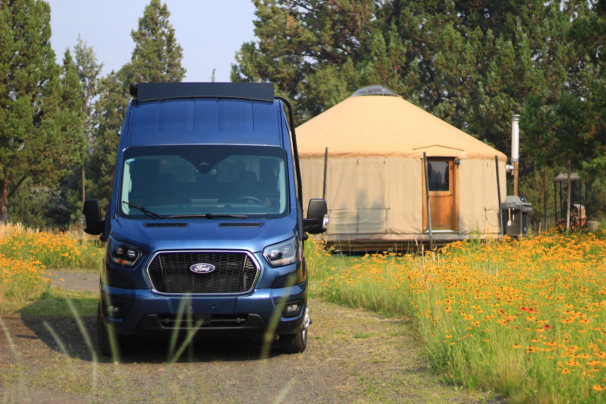 A blue Ford van parked on a dirt path in a field of yellow flowers, with a beige yurt and green trees in the background under a blue sky.