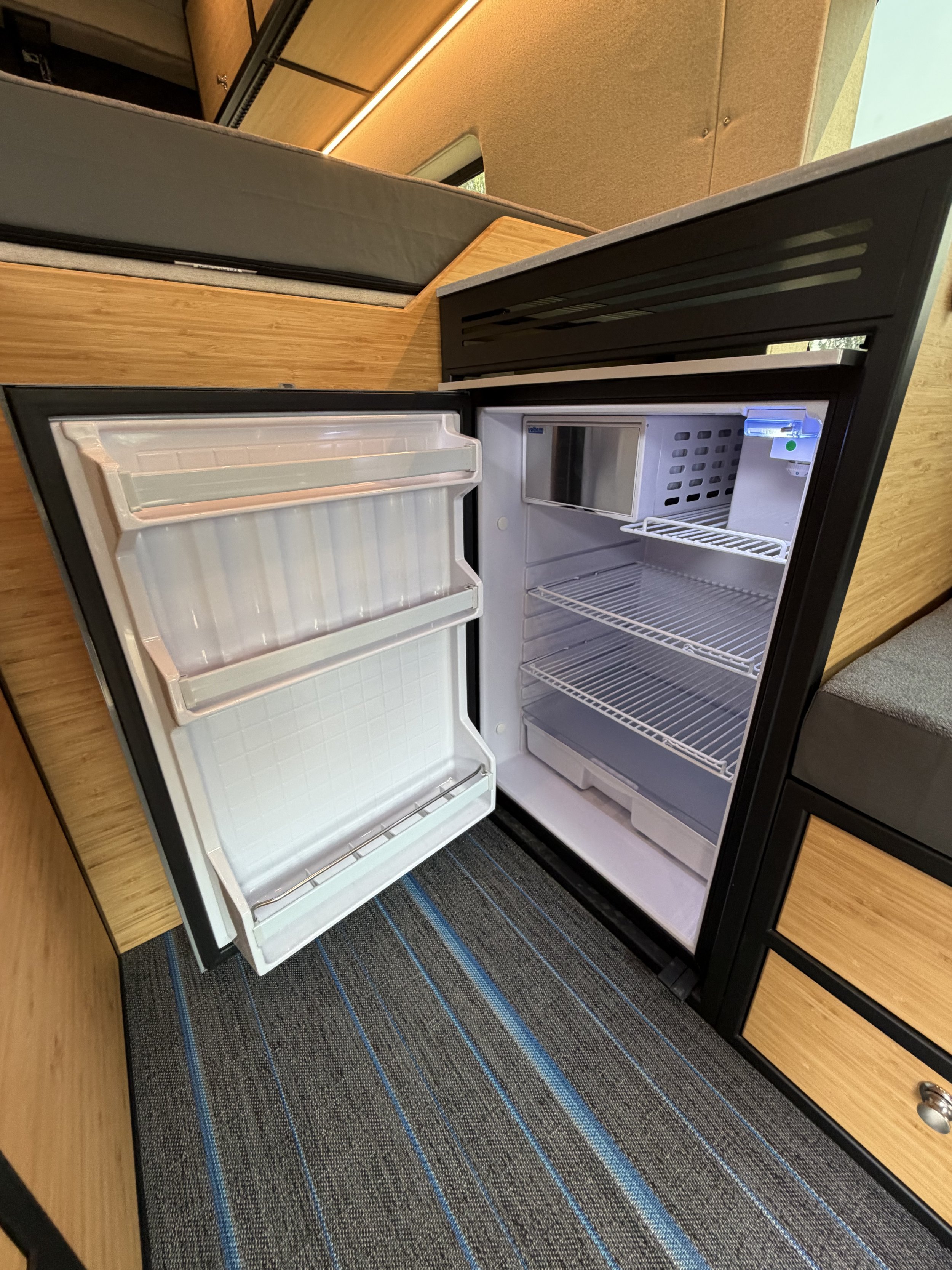 Open mini refrigerator with empty shelves on a wood and black cabinet. The floor is carpeted with a striped pattern.