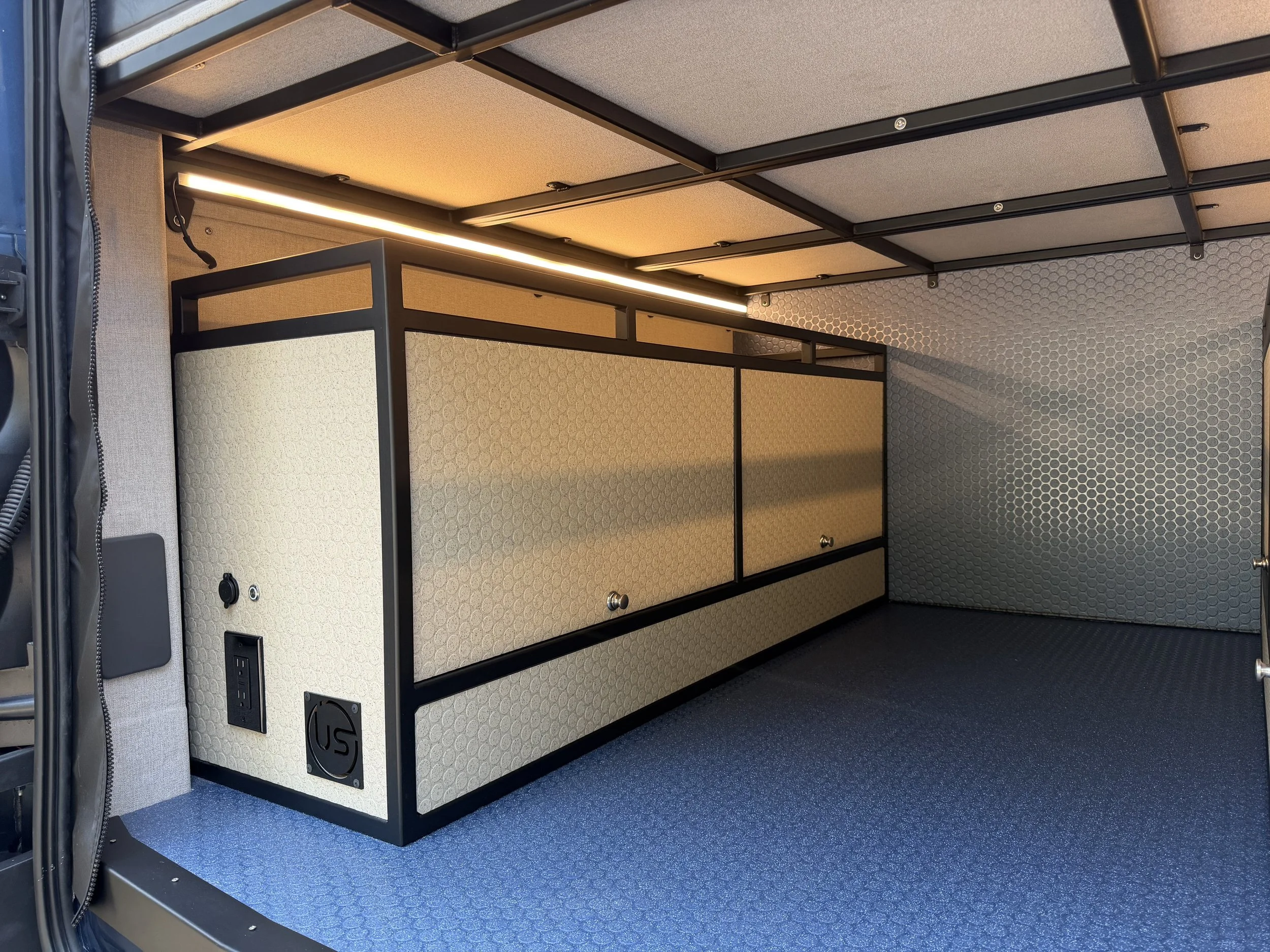 Interior of a small mobile room or office with beige walls, a black metal frame, and a strip of LED lighting along the ceiling. The floor is covered with dark blue textured material.